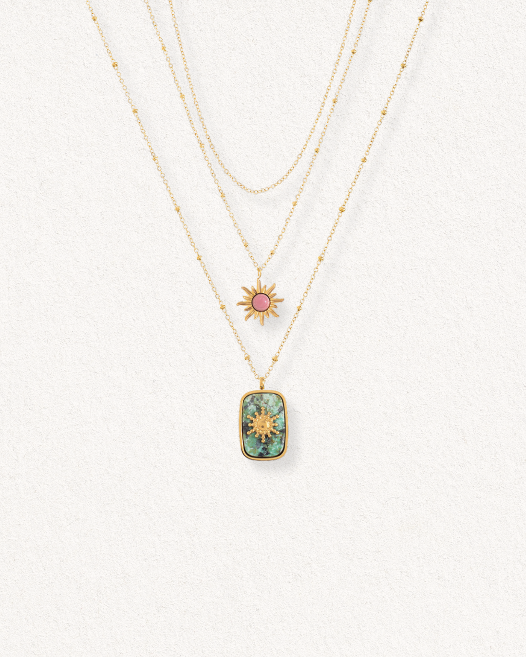 Kaklarota - Saule by Como Jewelry ir trīs zelta ķēdītes: vienkārša antialerģiska rotaslieta, rozā piekariņš ar saules stariem un tirkīza taisnstūra mūsdienīga dizaina kaklarota uz gaiša fona.