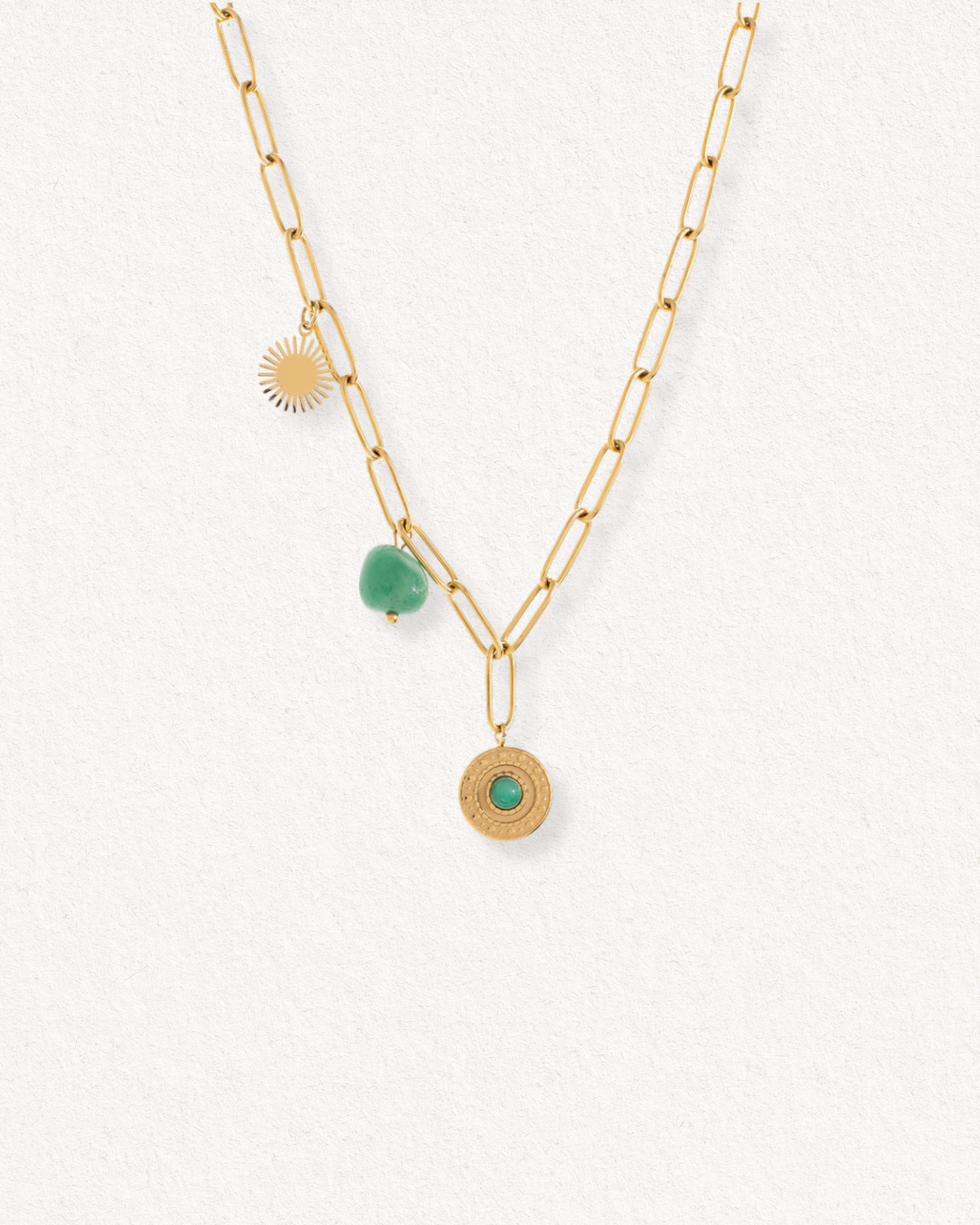 Como Jewelry 'Kaklarota - Zaļās saules ceļš': zelta kaklarota sievietēm ar trīs piekariņiem - saules staru, zaļu neregulāras formas krelli un noapaļotu medaļonu ar zaļu centru, izcelts uz balta teksturēta fona.