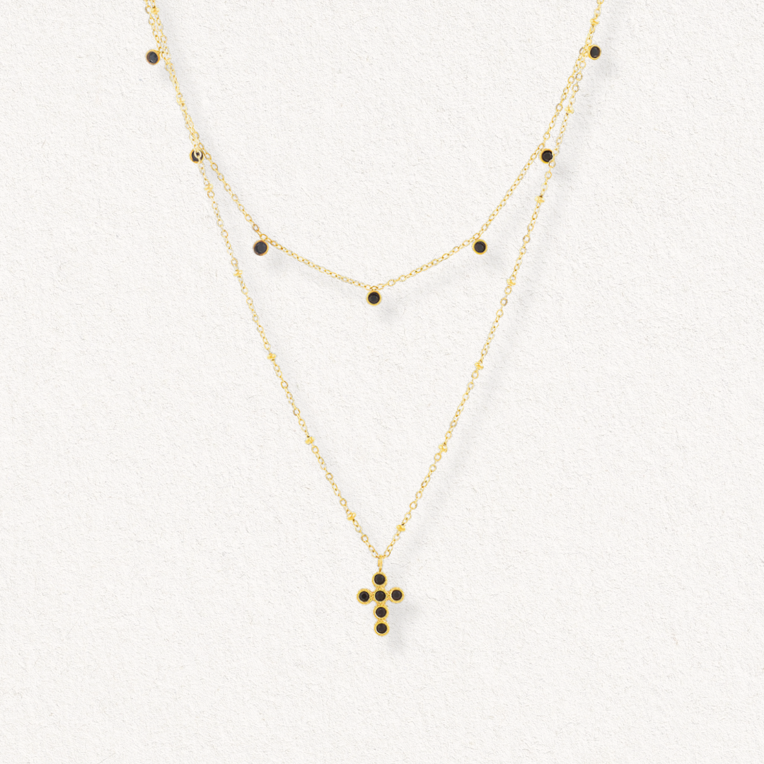 Como Jewelry Kaklarota - Mēness ēna ir zeltīta daudzslāņaina kaklarota ar divām maigām ķēdītēm: viena ar mazām melnām pērlītēm, otra ar melniem akmentiņiem rotātu krusta piekariņu. Šis antialerģiskais aksesuārs ir attēlots uz baltas faktūras virsmas.