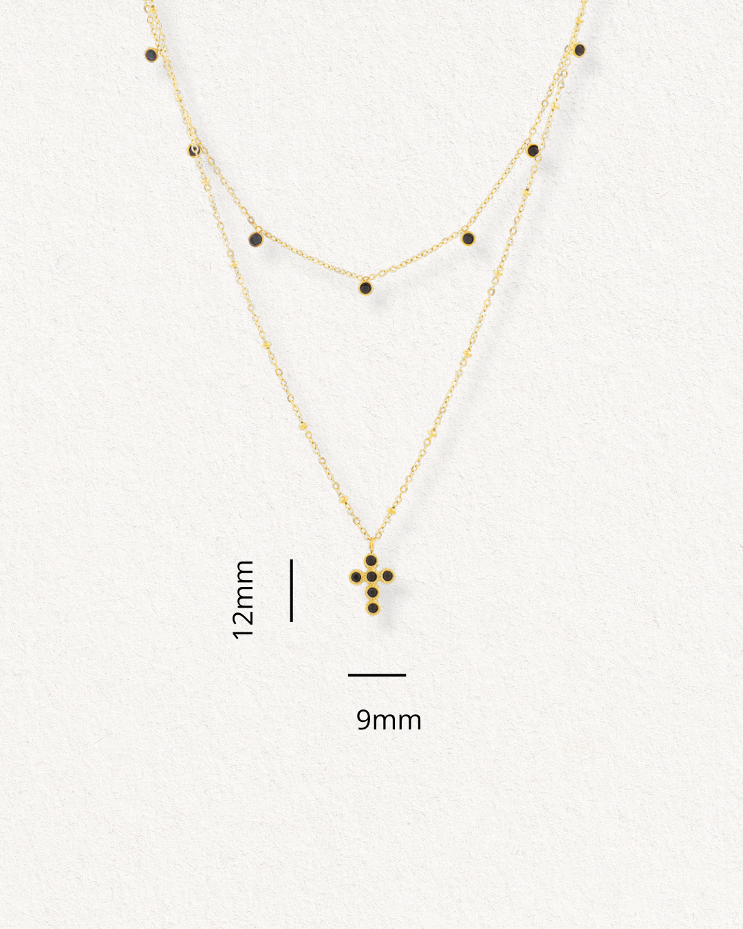 Kaklarota - Mēness ēna no Como Jewelry ir zeltīta kaklarota ar melniem akmeņu akcentiem; īsajā ķēdītē ir izvietoti akmeņi, bet garākajā - 9x12 mm liels krustiņš, kas attēlots uz gaiša fona.