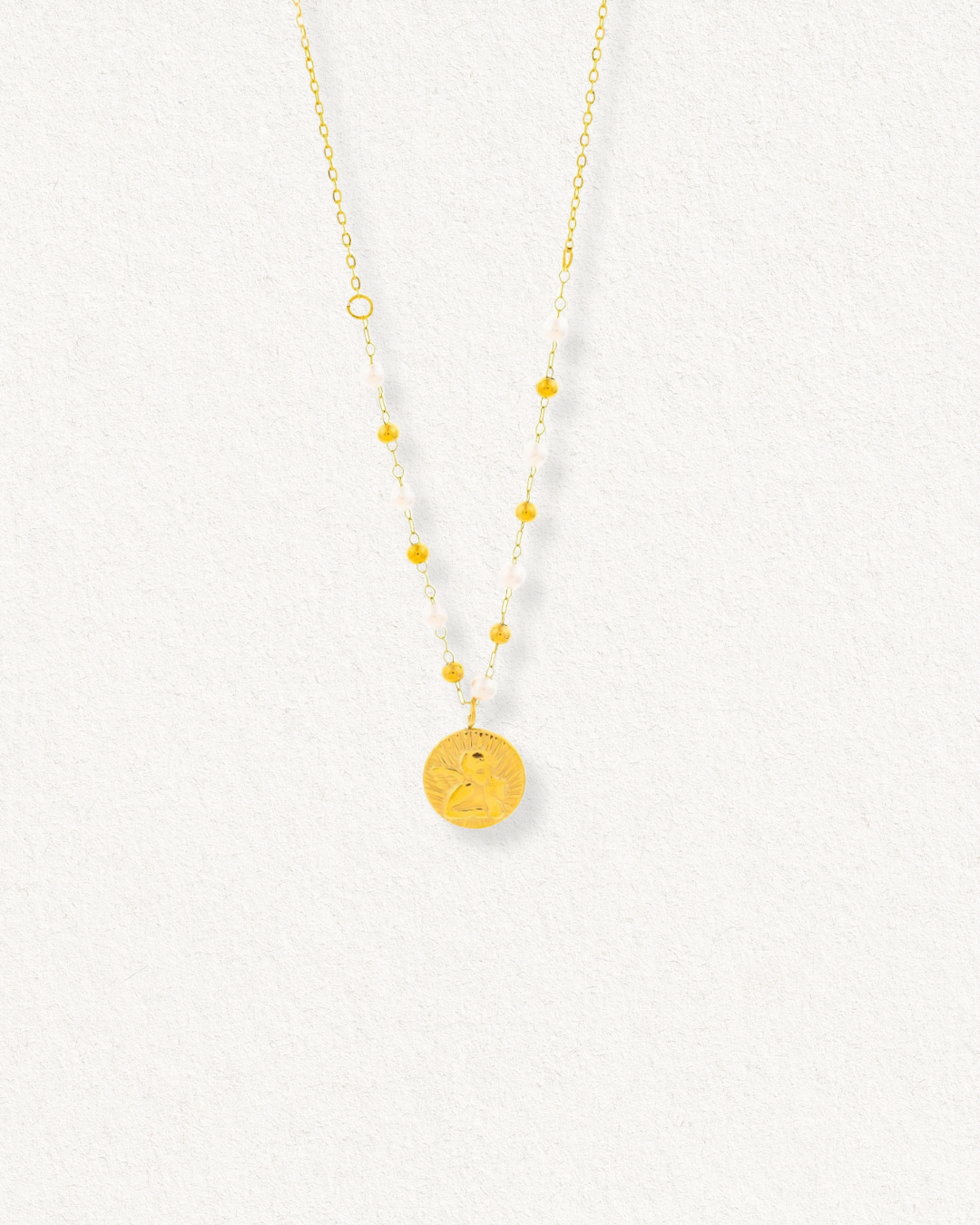 Kaklarota - Amors by Como Jewelry ir aprīkota ar plānu ķēdīti ar mazām zelta pērlītēm un apaļu, iegravētu kulonu. Šis modernais izstrādājums, kas izvietots uz gaišas faktūras fona, jebkuram tērpam piešķir pārlaicīgu eleganci.