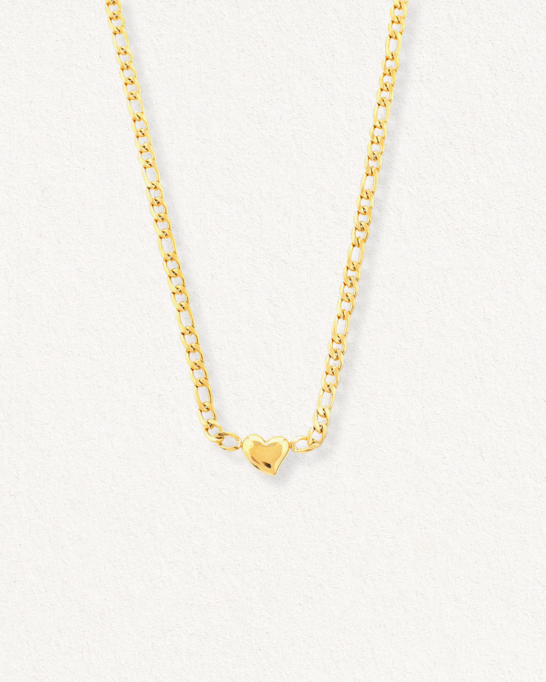 Kaklarota - Sirds by Como Jewelry ir zelta kaklarota ar nelielu, spīdīgu sirds formas piekariņu centrā uz gaiša, teksturēta fona.