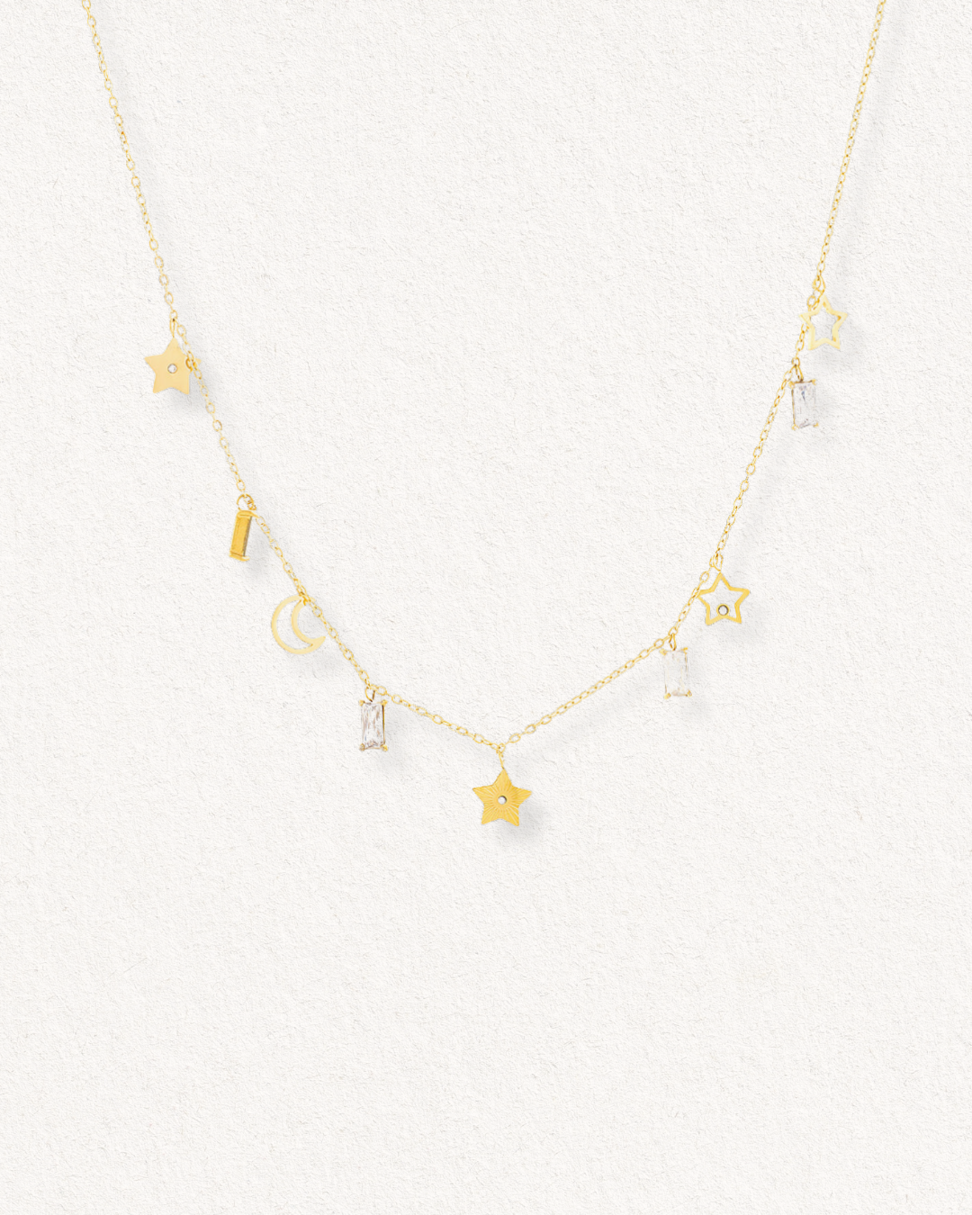 Kaklarota - Zvaigžņu deja no Como Jewelry ir maiga Juveliertērauda kaklarota ar maziem piekārtiem piekariņiem - zvaigznēm, taisnstūriem, pusmēnesi un caurspīdīgiem dārgakmeņiem, kas izvietoti uz viegla faktūras fona.