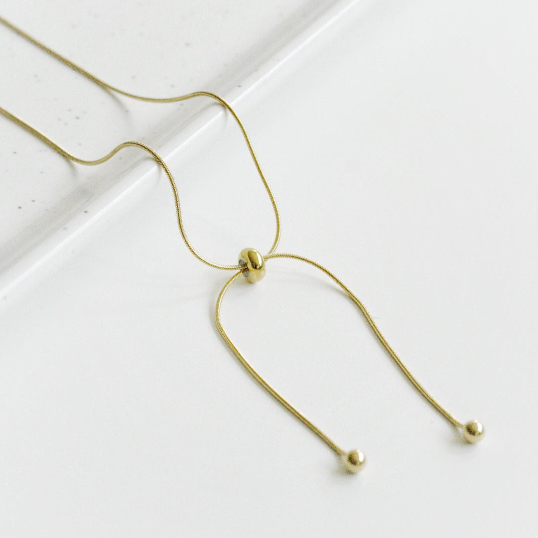 Kaklarota - Zelta cilpa no Como Jewelry ir maiga, apzeltīta kaklarota ar bīdāmu pērlīšu aizdares aizdari un diviem galiem, no kuriem katru rotā maza zelta pērlīte, kas izvietota uz baltas virsmas.