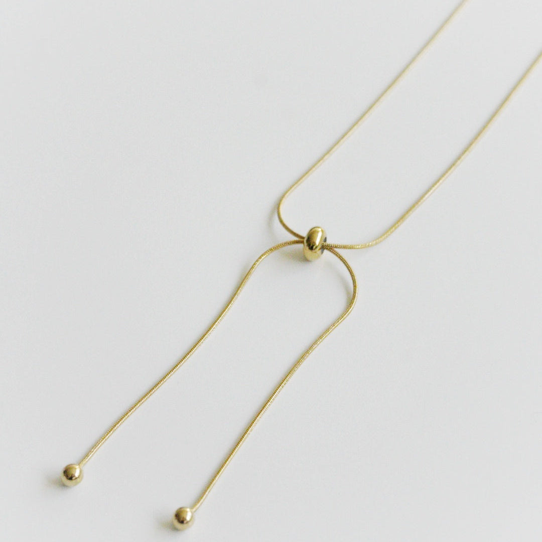 Como Jewelry Kaklarota - Zelta cilpa ir maiga, apzeltīta kaklarota ar bīdāmu pērlīti un diviem ķēdītes galiem, no kuriem katru rotā maza zelta lodīte, kas izvietota uz balta fona.