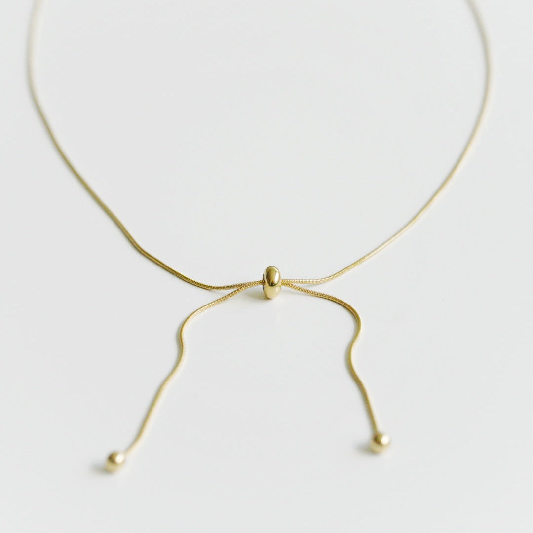 Como Jewelry Kaklarota - Zelta cilpa: maiga, apzeltīta kaklarota ar regulējamu bīdāmu pērlīti un diviem vaļējiem ķēdītes galiem ar mazām zelta pērlītēm. Uz balta fona. Mitrumizturīga, lai nodrošinātu noturīgu spīdumu.