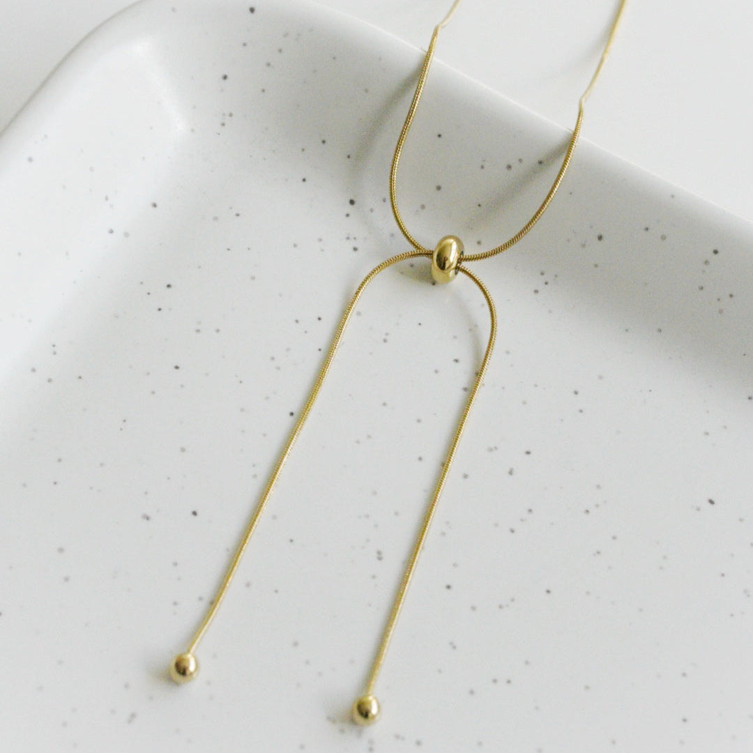 Como Jewelry Kaklarota - Zelta cilpa: Kārtīga zeltīta kaklarota ar bīdāmu pērlīti un diviem karināmiem galiem, katrs no kuriem noslēdzas ar mazu zelta bumbiņu. Uz balta plankumaina paliktņa. Mitrumizturīga un piemērota ikdienas valkāšanai.