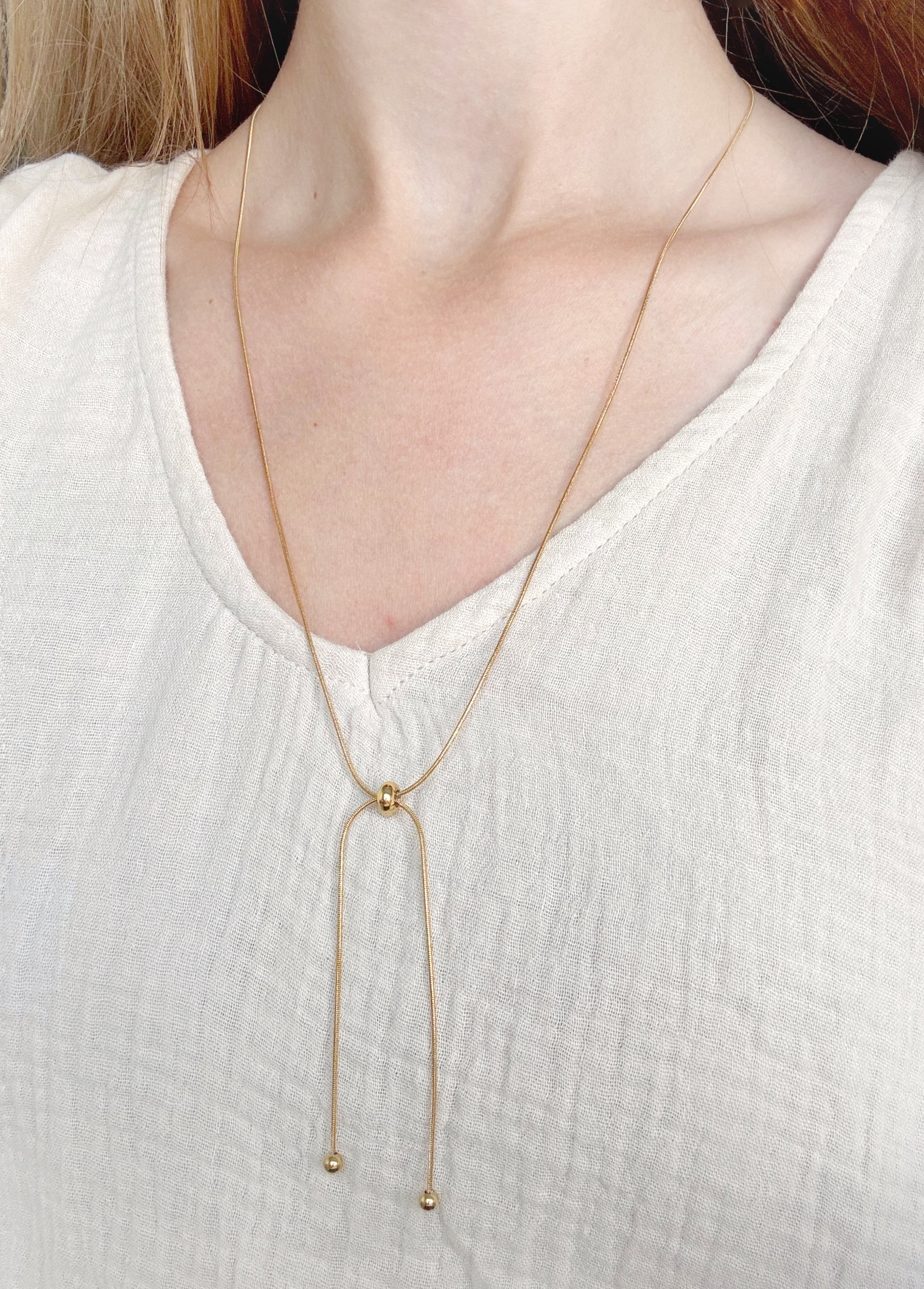 Persona baltā fakturētā topā modelē Como Jewelry Kaklarota - Zelta cilpa, nerūsējošā tērauda lariat kaklarotu ar centrālo gredzenu un diviem zelta pērlīšu galiem, padarot to noturīgu pret mitrumu.