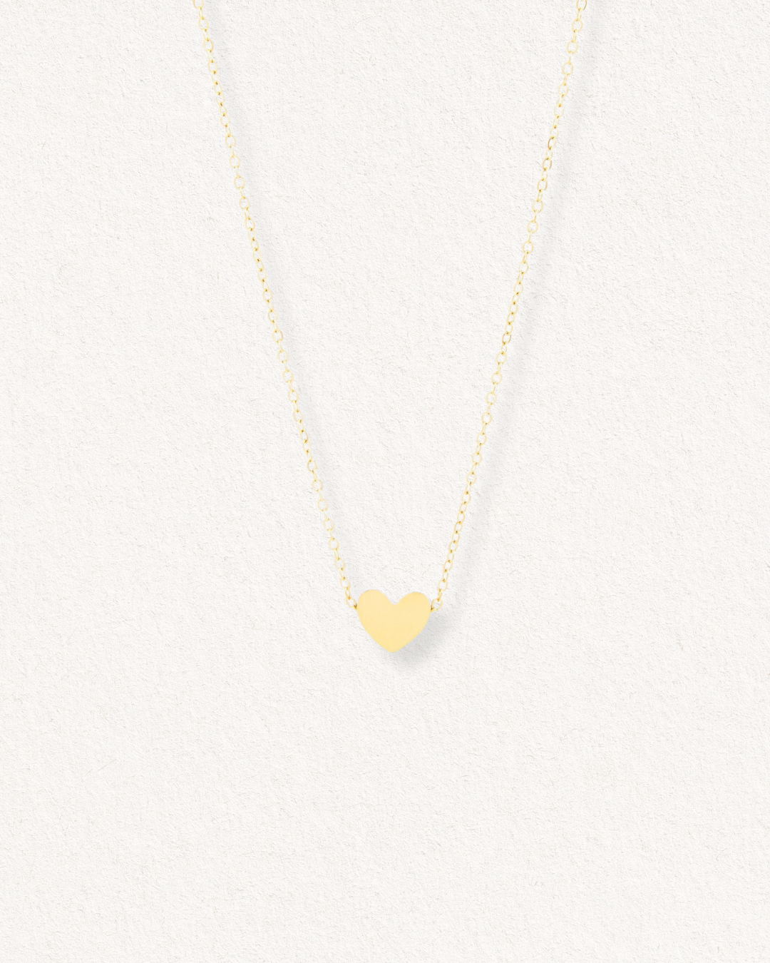 Kaklarota - Sirds by Como Jewelry ir neliela sirds piekariņš uz smalkas zelta ķēdītes, kas eleganti izvietots uz gaišas faktūras fona.