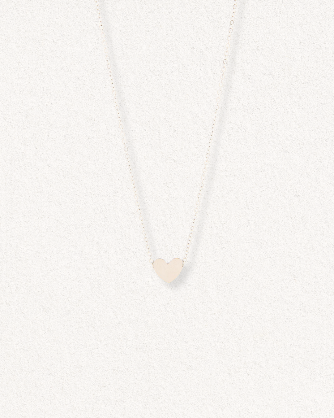 Kaklarota - Sirds by Como Jewelry ir neliels, plakans sirds formas kuloniņš uz smalkas ķēdītes, kas eleganti izvietots uz gaišas faktūras fona.