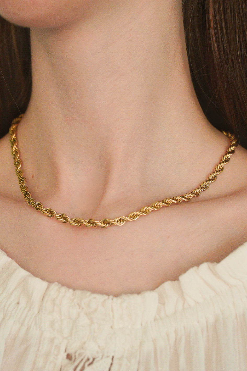 Tuvplānā redzama sieviete, kas valkā Como Jewelry Kaklarota - Zelta ķēde, vītā zelta virves ķēdīte, ar baltas faktūras blūzi. Uzmanības centrā ir kaklarota pret viņas kailo kaklu un kakla kaulu.