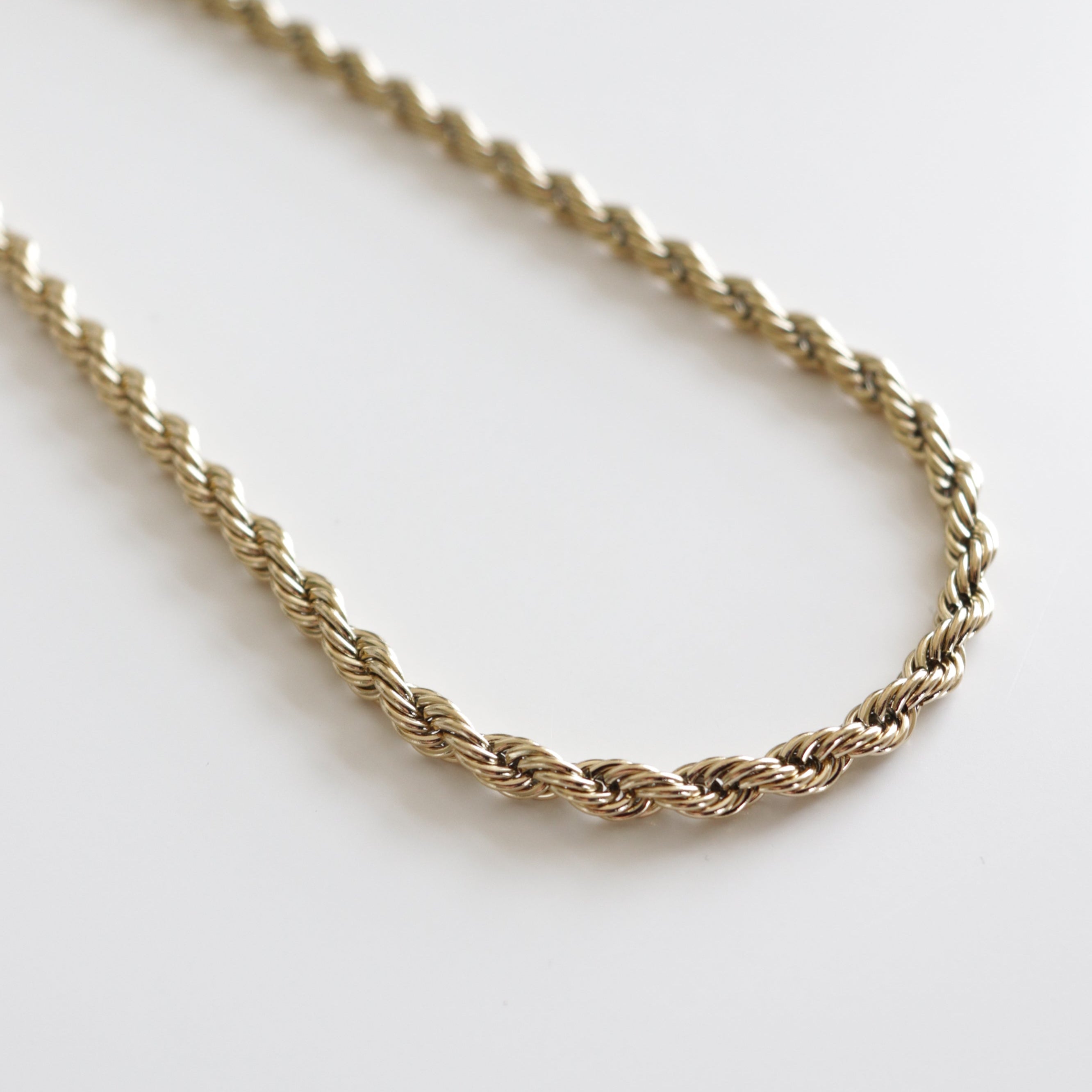 Kaklarota - Zelta ķēde no Como Jewelry ar modernu dizainu un zelta virves ķēdīti, kas izvietota uz vienkrāsainas baltas virsmas.