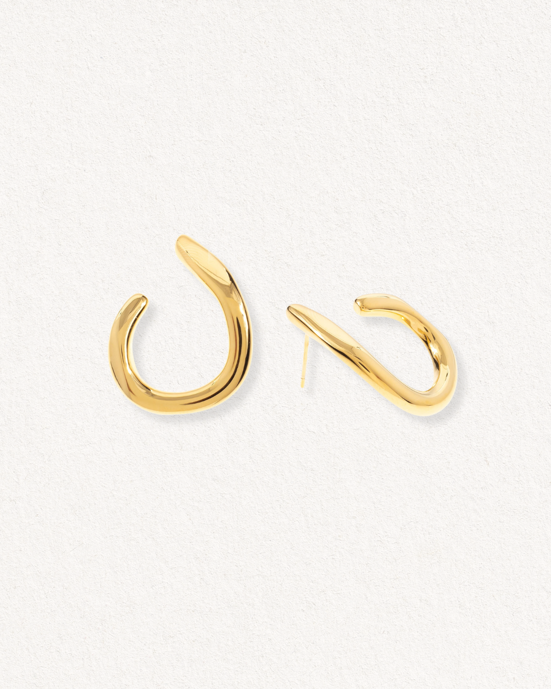 Auskari - Plūsma by Como Jewelry: zelta riņķveida auskari ar abstraktu, neregulāru, atvērtu formu uz gaiši bēša faktūras fona.