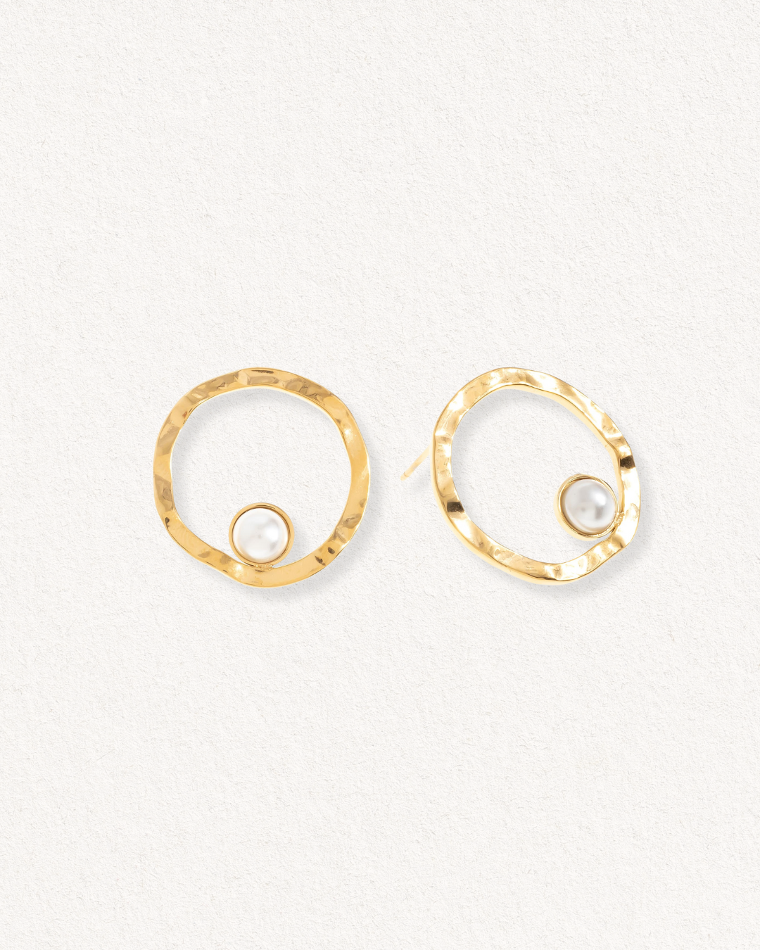 Como Jewelry "Mazie auskari - Rietošā pērle" ir neregulāras apzeltītas atklātas cilpas, katra no tām apakšā akcentēta ar nelielu baltu pērli, kas izvietota uz gaišas faktūras fona, lai radītu mūsdienīgu noskaņu.