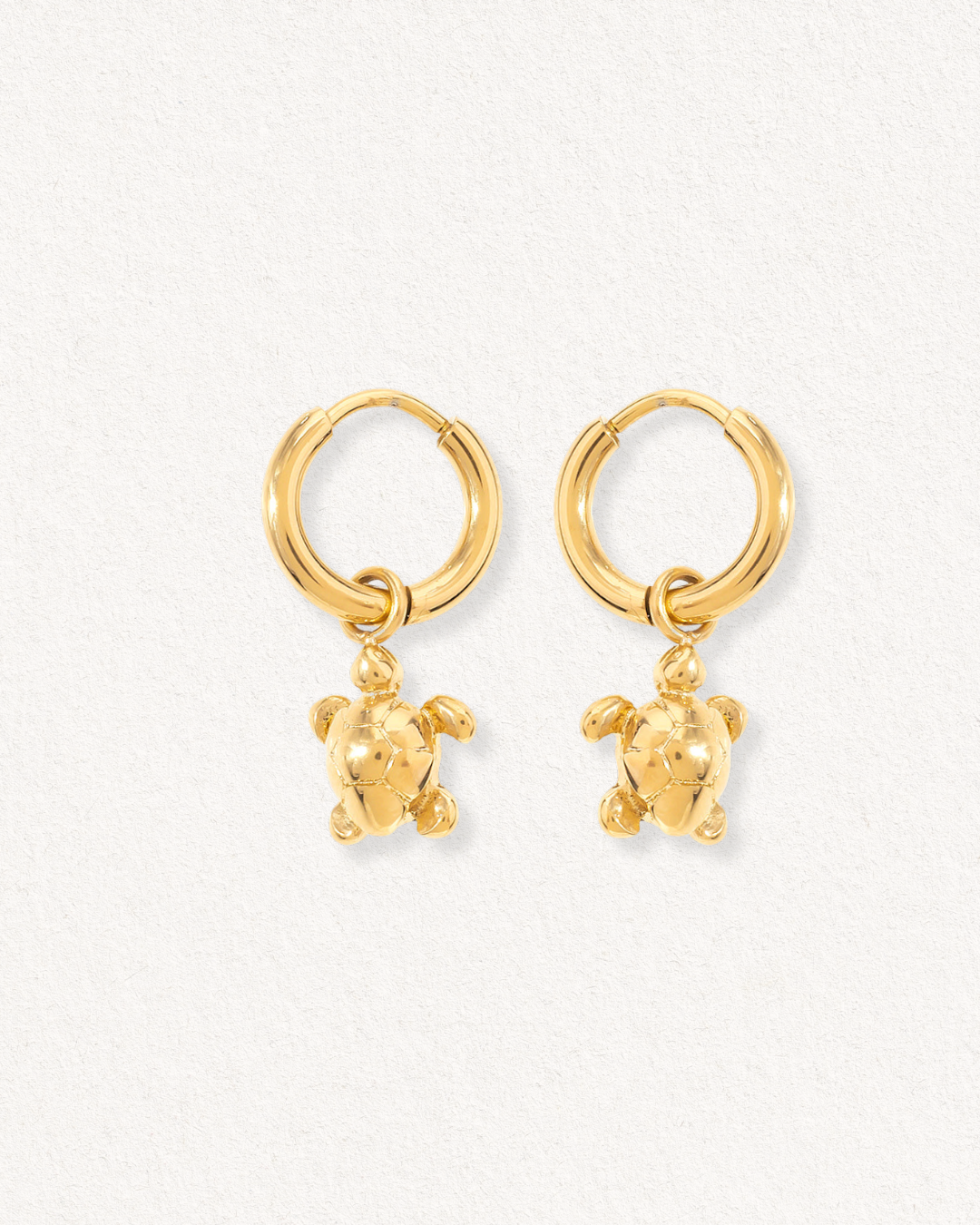 Auskari - Miers: zeltīti auskari ar smalkiem bruņurupuču piekariņiem, kas karājas no katra riņķa, izvietoti uz gaiša, teksturēta fona.