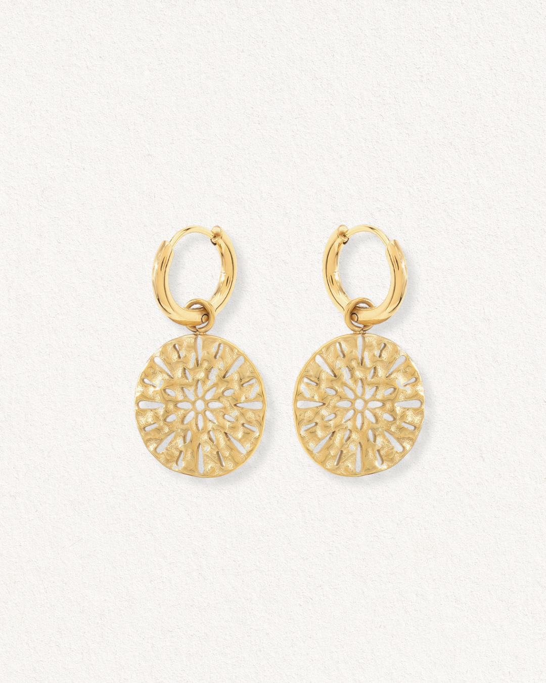 Auskari - Saules raksti no Como Jewelry: zelta riņķveida auskari ar apaļiem, sarežģītiem ziedu piekariņiem uz gaišas faktūras fona. Moderni auskari ikdienai un piemēroti jutīgām ausīm.