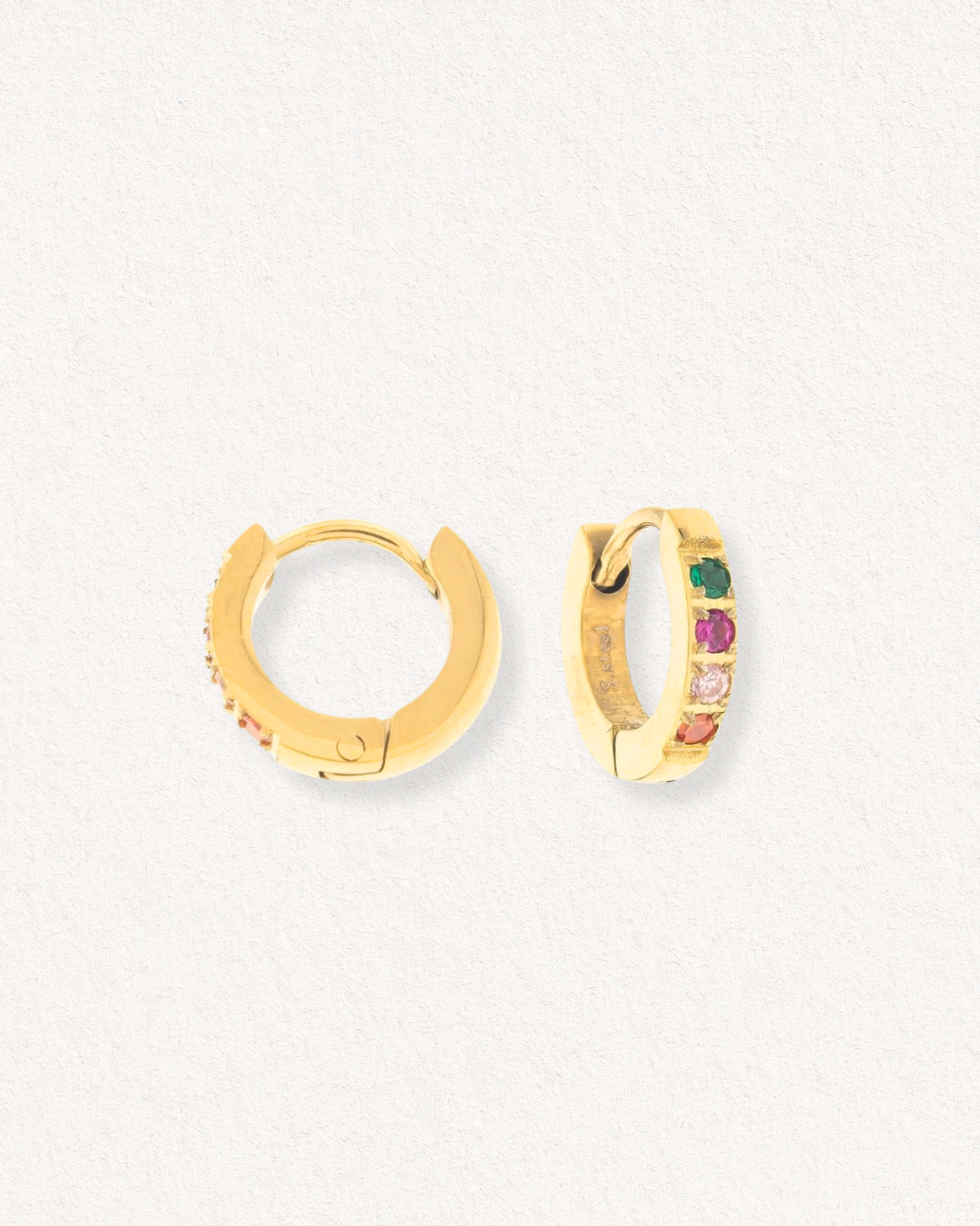 Uz gaiša, fakturēta fona ir izvietoti divi Como Jewelry "Mazie auskari - Krāsainie kristāli" - nelieli tērauda auskari ar zeltītu pārklājumu, kurus rotā krāsainu zaļu, rozā un caurspīdīgu kristālu rinda gar priekšpusi.