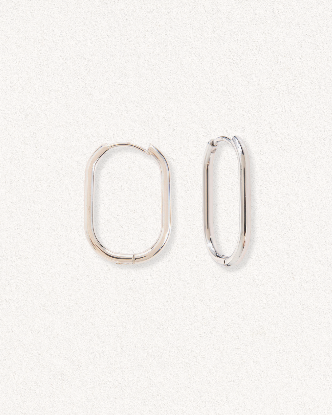 Divi Mazie auskari - Ovāls by Como Jewelry, kas izgatavoti no nerūsējošais tērauds, ir attēloti viens otram blakus uz gaiša fakturēta fona. Viens auskars ir nedaudz atvērts, bet otrs ir aizvērts, veidojot mūsdienīgu izskatu.