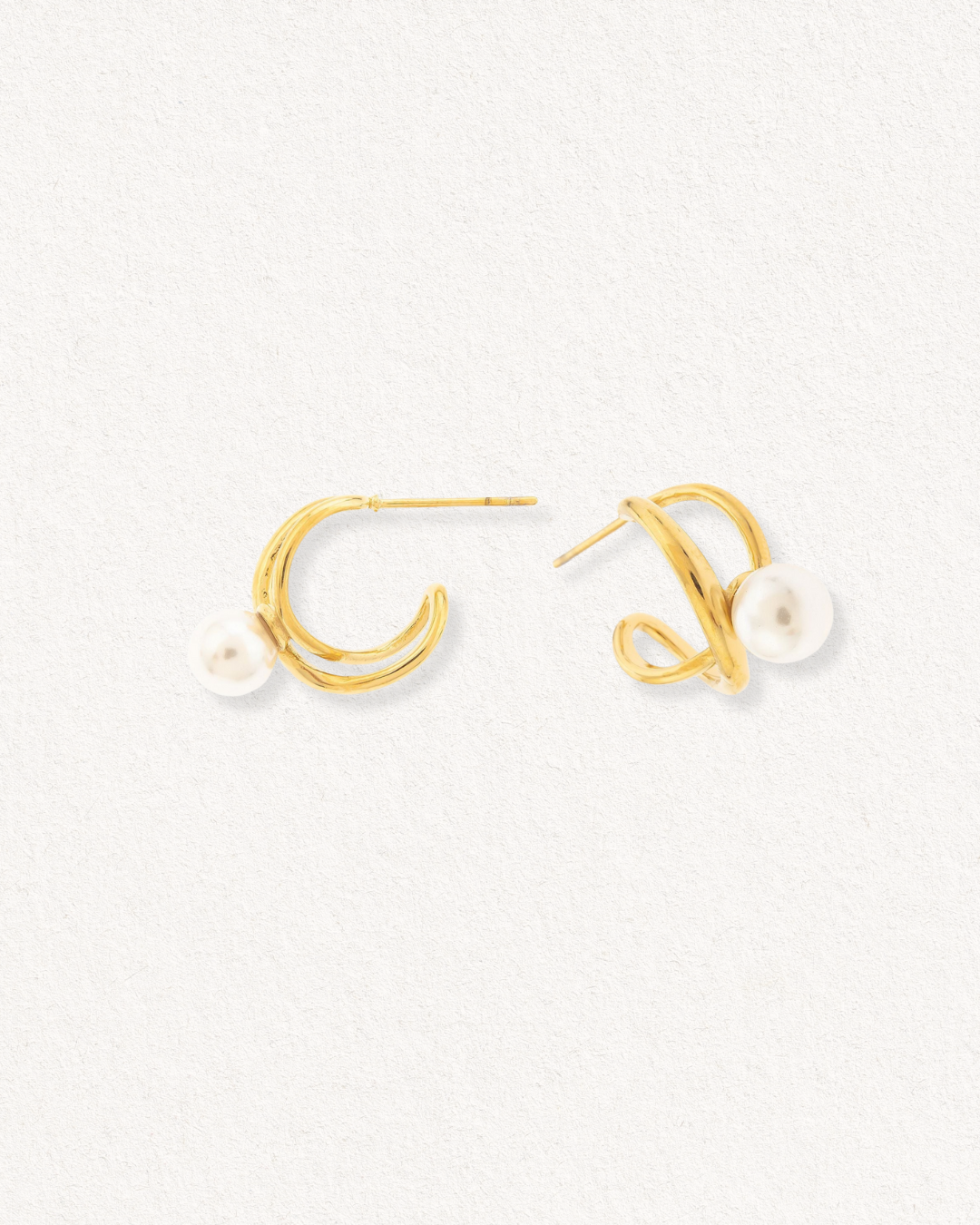 Mazie auskari - Loop by Como Jewelry ir zelta auskari ar abstraktu, virpuļveidīgu dizainu un vienu imitācijas pērli uz katra. Attēlots uz gaiša, teksturēta fona.