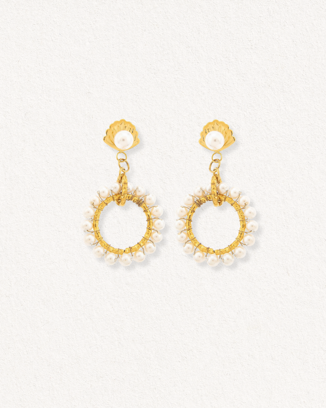 Como Jewelry Mazie auskari - Pērļu dārzs: zelta cilpas, kas rotātas ar mazām perlamutra pērlītēm gar malām, katra piekārta pie zelta gliemežnīcas formas tapas ar pērļu centru, izvietota uz gaišas faktūras fona.