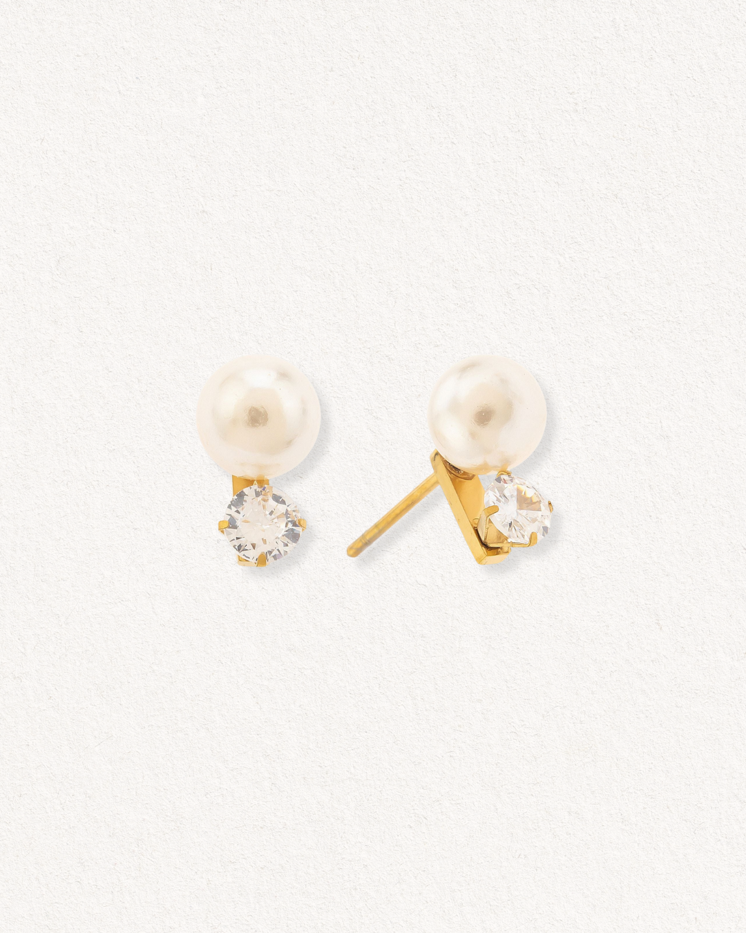 Mazie auskari - Klusā gaisma by Como Jewelry: zeltīti minimālisma auskari ar apaļu baltu pērles imitāciju virs caurspīdīga apaļa dārgakmens, kas attēlots uz gaišas faktūras fona.