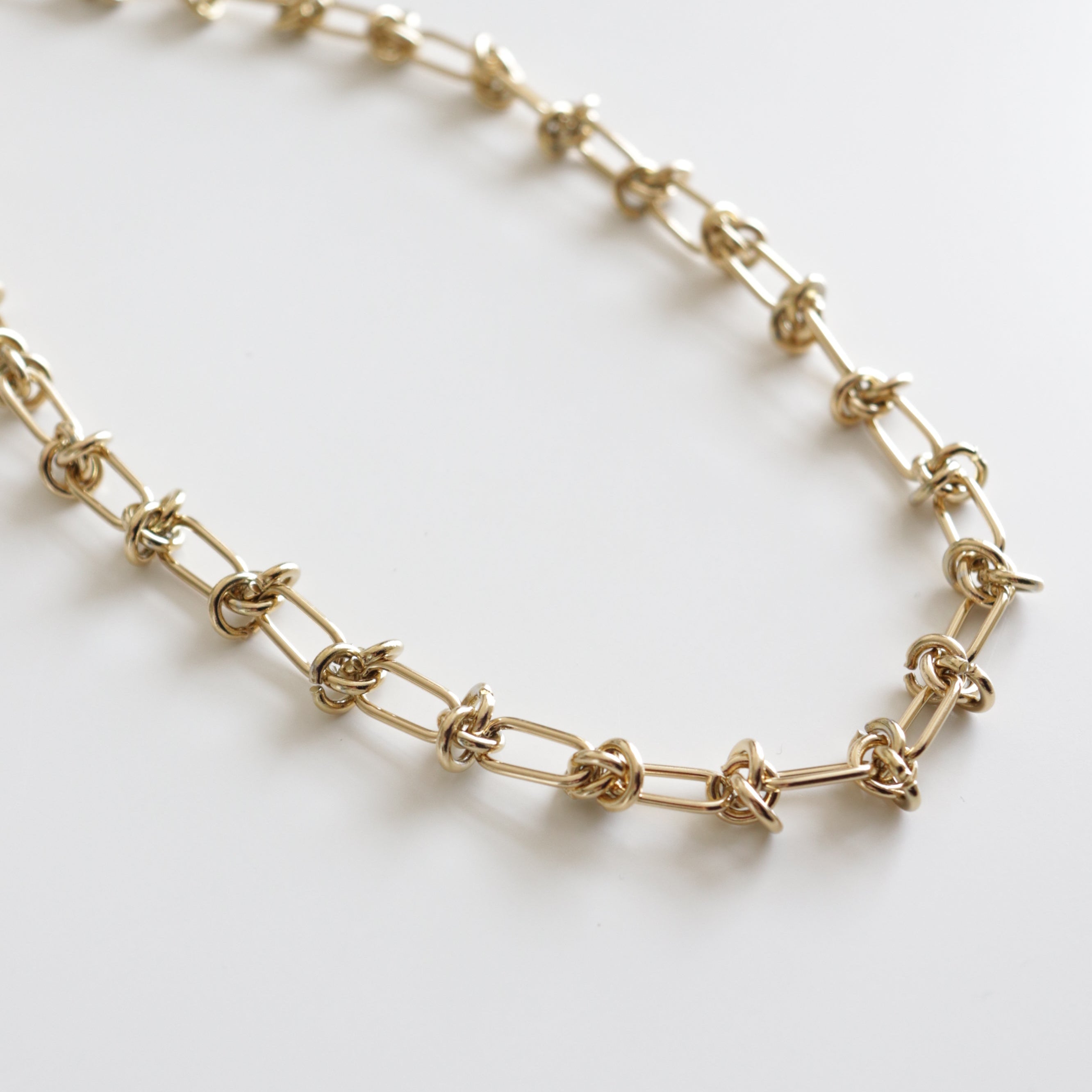 Como Jewelry Kaklarota - Mezglu spēks ir veidots no zeltītiem savienojumiem ar elegantām mezglu detaļām uz balta fona.