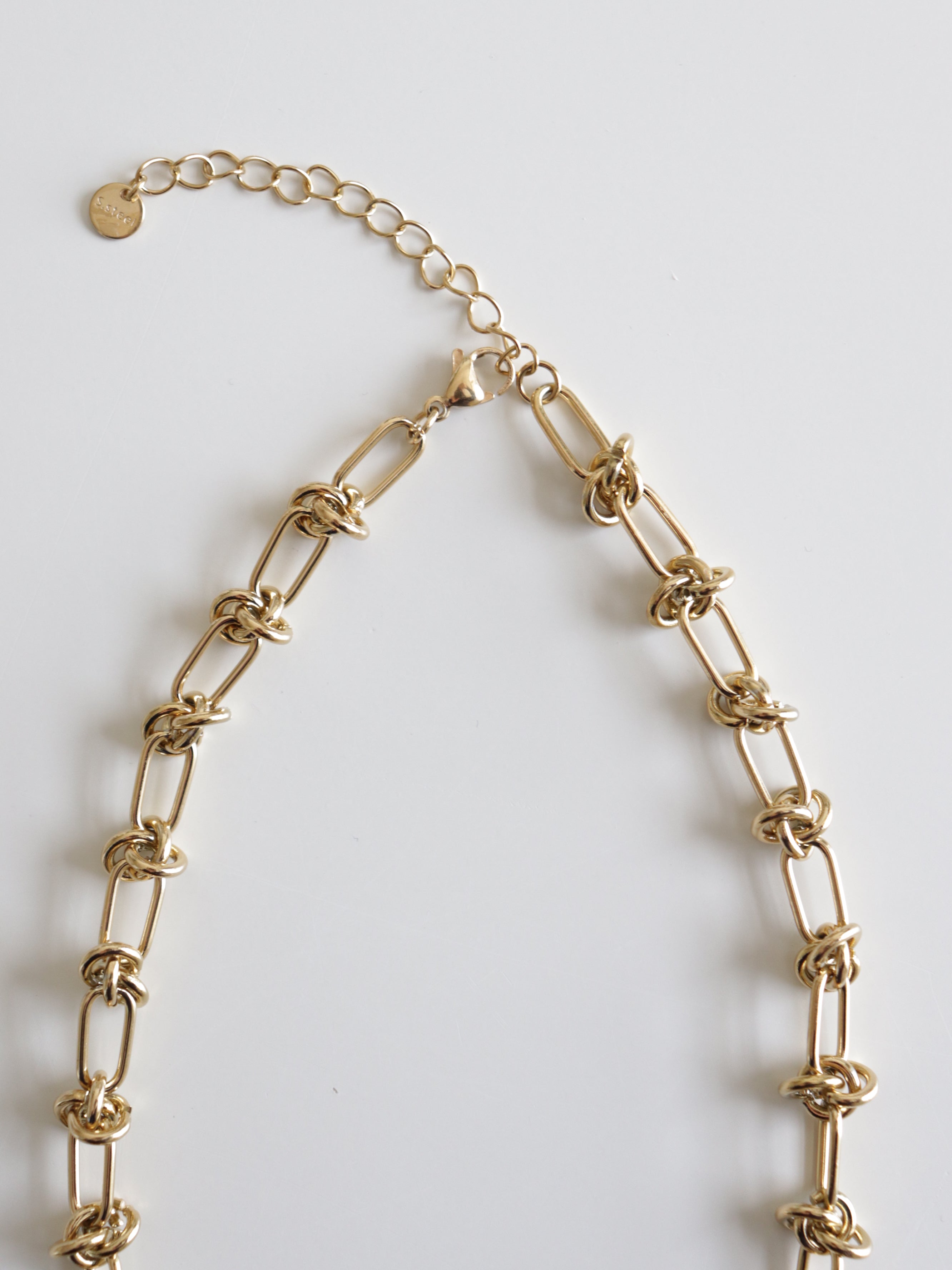 Kaklarota - Mezglu spēks no Como Jewelry, juveliertērauda kaklarota ar taisnstūrveida posmiem, mezglu detaļām, regulējamu pagarinājuma ķēdīti un mazu apaļu birku, kas attēlota uz balta fona.