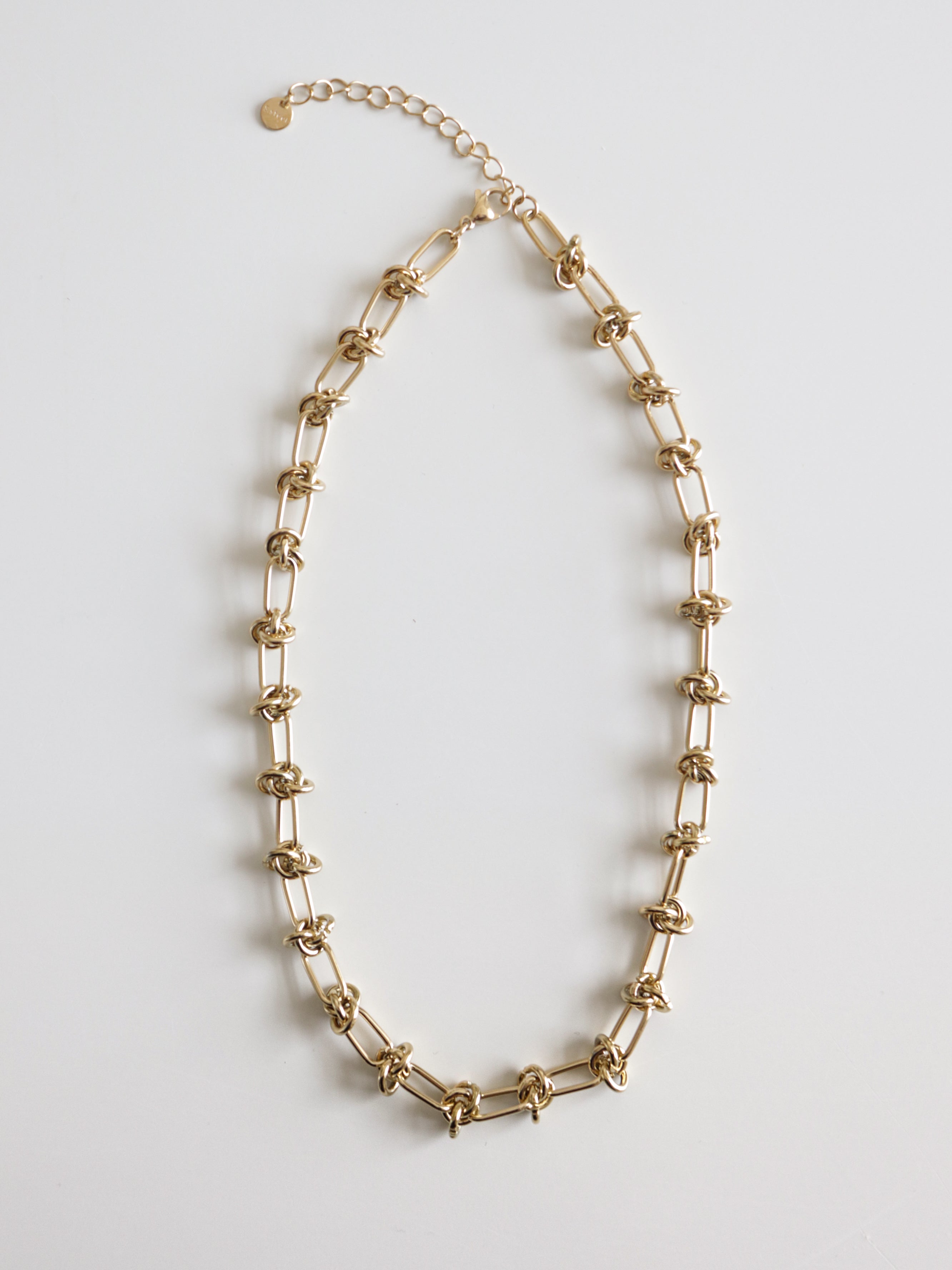 Como Jewelry "Kaklarota - Mezglu spēks" - moderna zelta kaklarota sievietēm ar taisnstūra posmiem un mezgliņiem, regulējama aizdare ar mazu apaļu piekariņu. Izklāta uz balta fona.