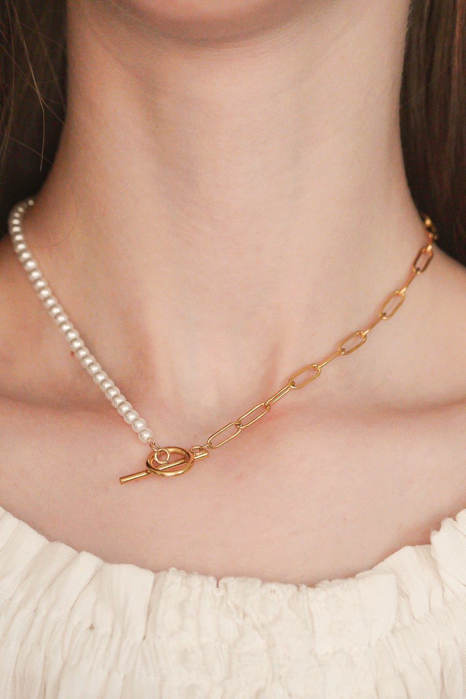 Sievietes kakla tuvplānā redzams Como Jewelry Kaklarota - ar pērlēm, mūsdienīga dizaina kaklarota ar pusi baltām pērlēm, pusi zelta ķēdes posmiem un zelta aizdares aizdari. Šī antialerģiska rotaslieta papildina viņas krēmkrāsas topu.