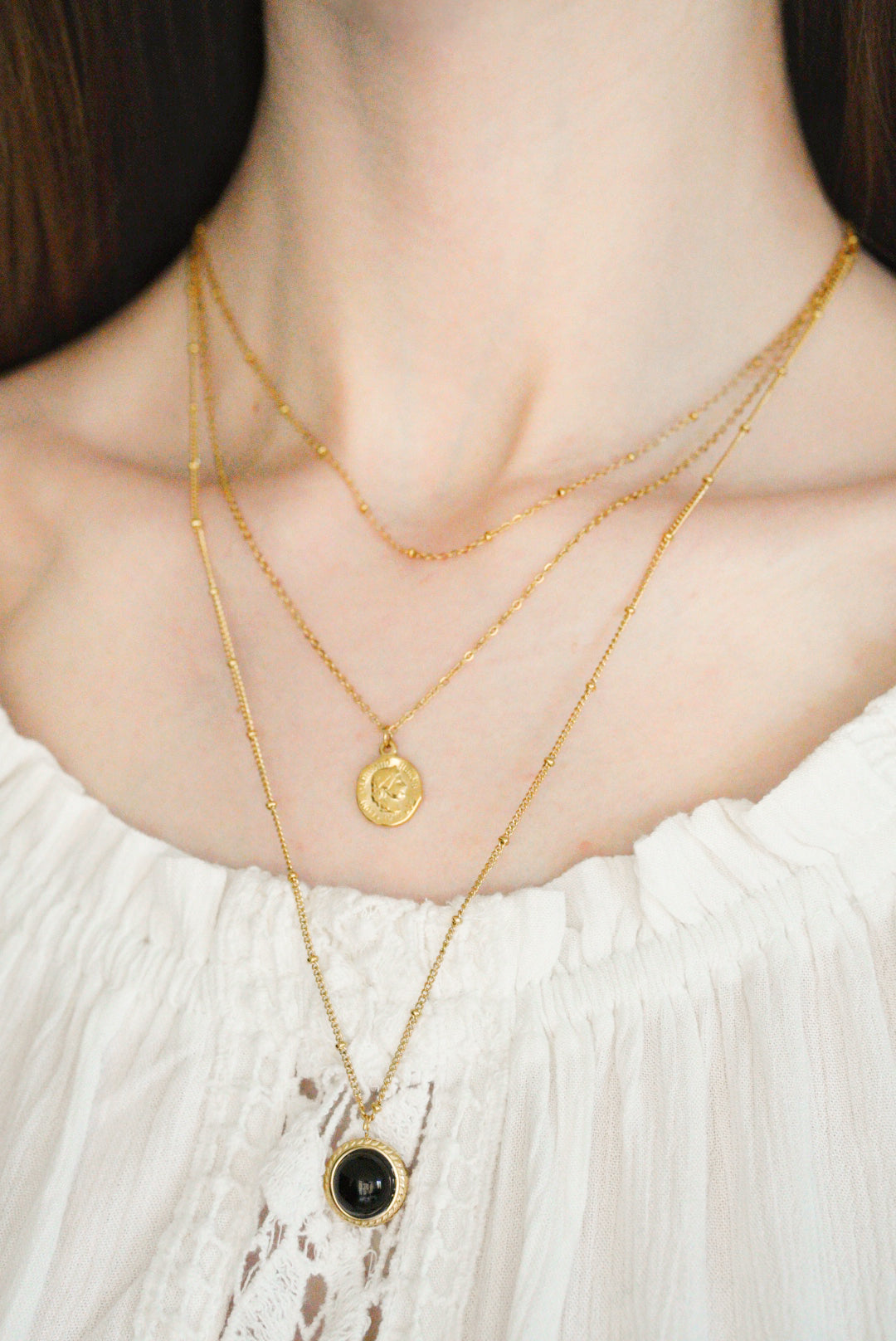 Sieviete baltā topā bez pleca demonstrē trīs Como Jewelry Kaklarota - Nakts simboli, tostarp antialerģiska rotaslieta ar mazu apaļu piekariņu un viena ar melnu akmens piekariņu.