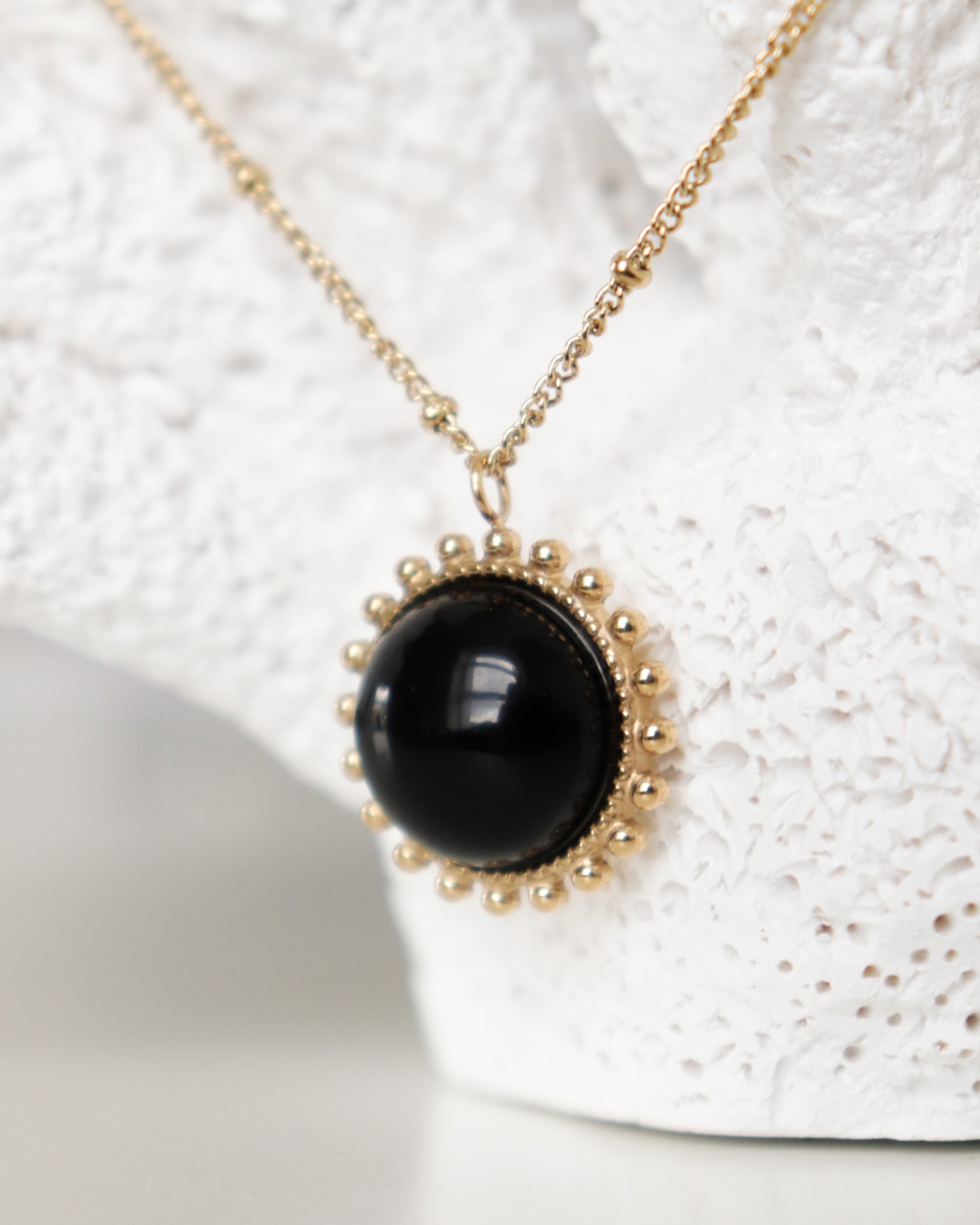 Mūsdienīga Como Jewelry zelta kaklarota - Saules akmens ar apaļu melnu piekariņu un smalkām zelta bumbiņām ir izstādīta uz teksturēta balta fona.