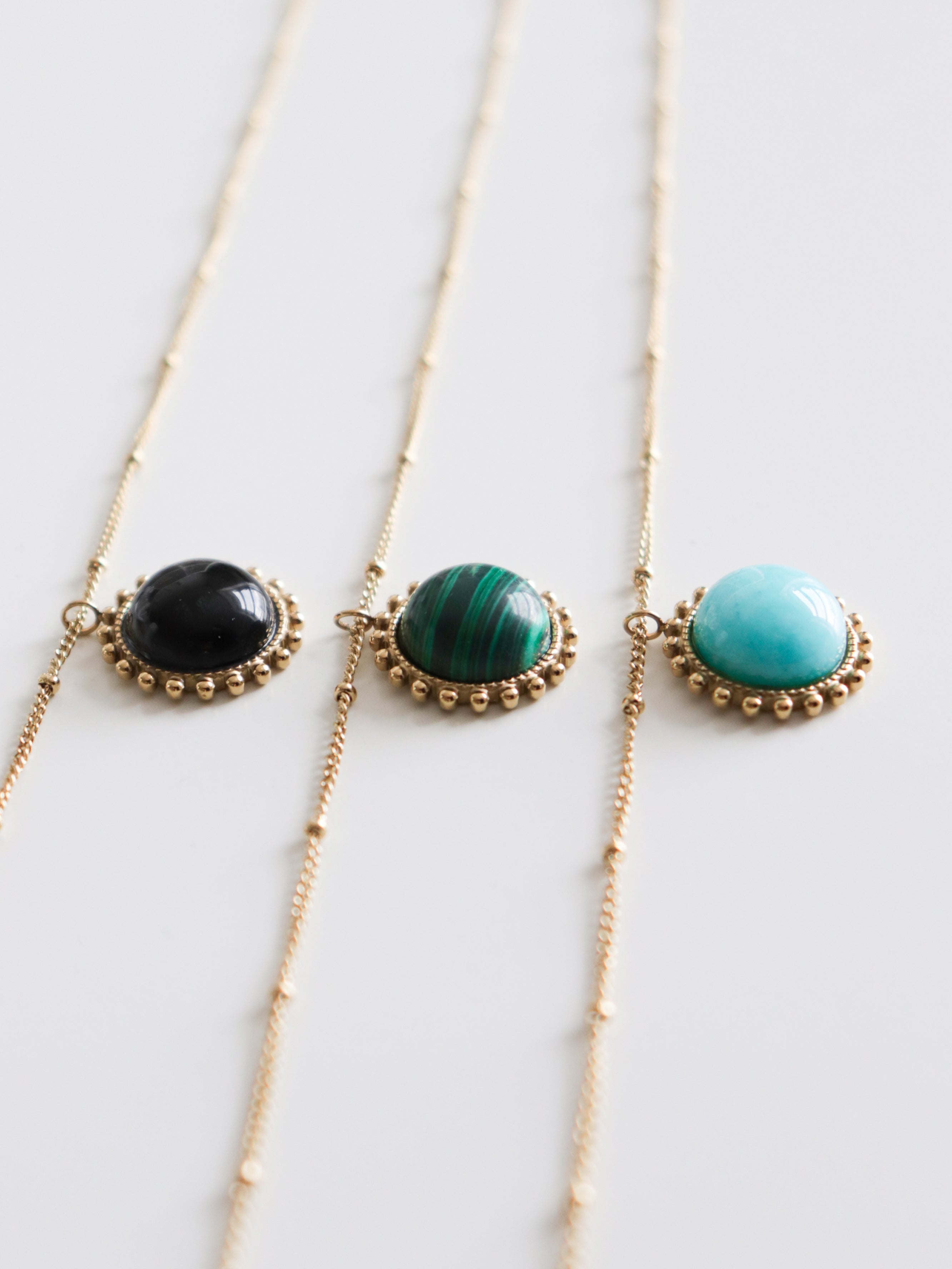 Trīs zelta ķēdīšu kaklarotas no Como Jewelry, katra Kaklarota - Saules akmens, ar apaļiem piekariņiem no pulētiem melniem, zaļi svītrainiem un tirkīza ziliem akmeņiem, kas eleganti izkārtoti uz baltas virsmas.