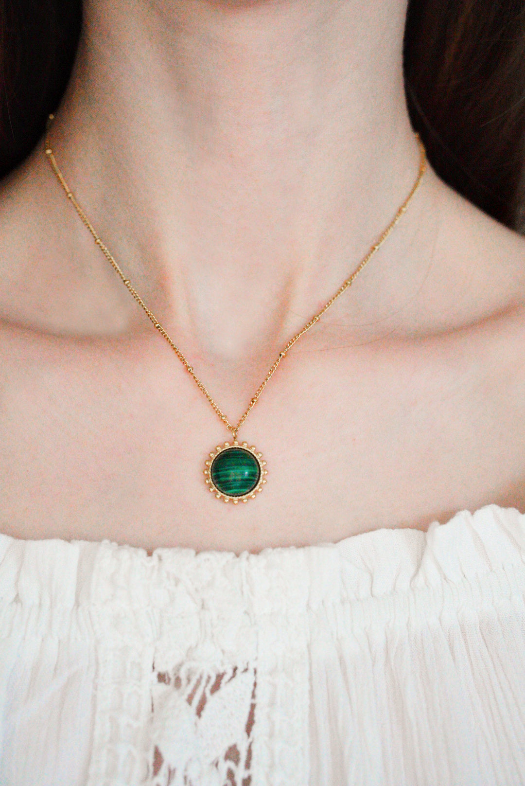 Persona, kas valkā Como Jewelry Kaklarota - Saules akmens ar apaļu zaļu piekariņu, tērpusies baltā, teksturētā, no pleca atdalītā topā.