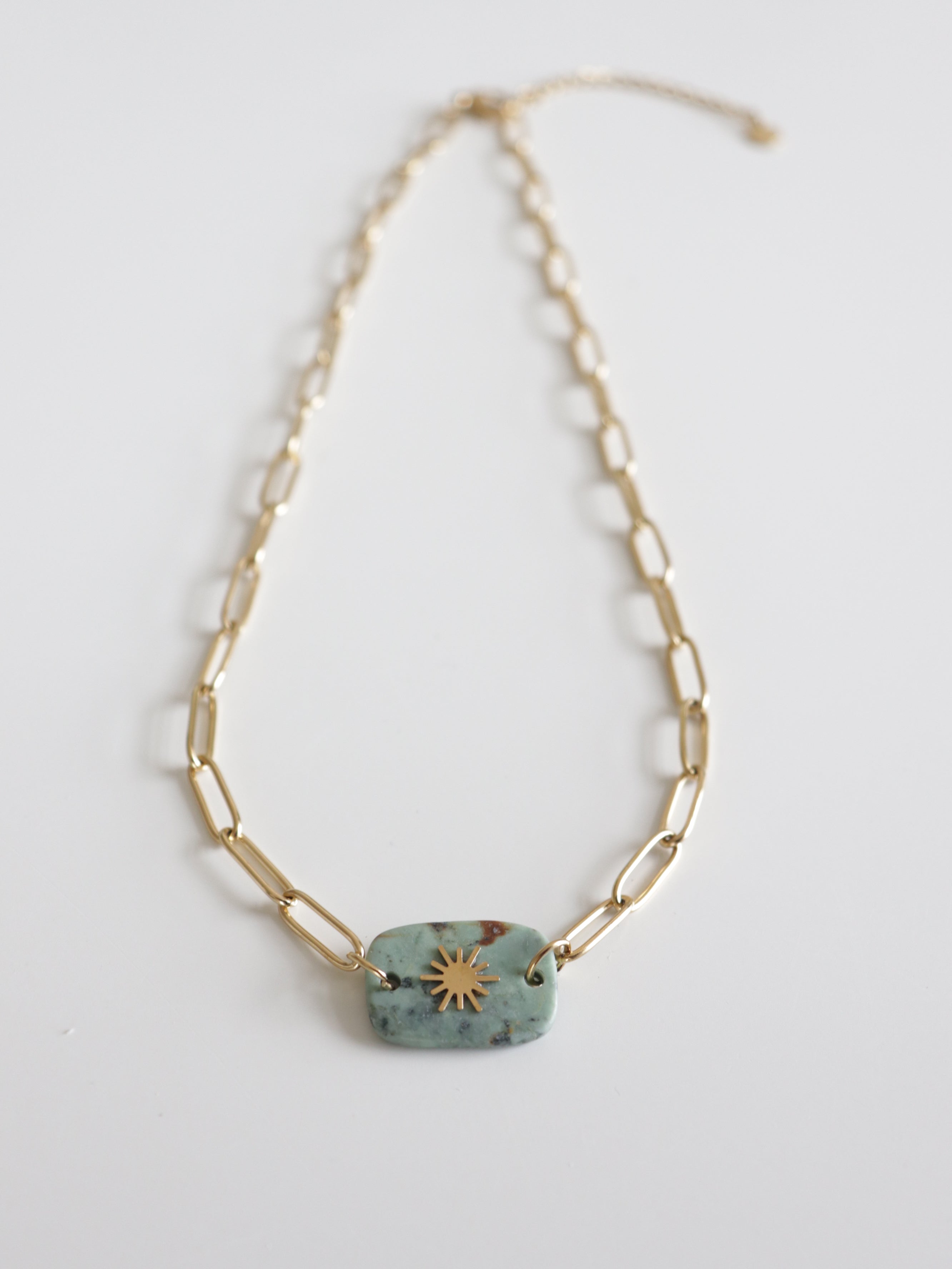 Kaklarota - Saules akmens ceļš by Como Jewelry ir sieviešu zelta kaklarota ar taisnstūra formas posmiem un zaļa akmens piekariņu, kura centrā attēlots zelta saules stariņš uz balta fona.