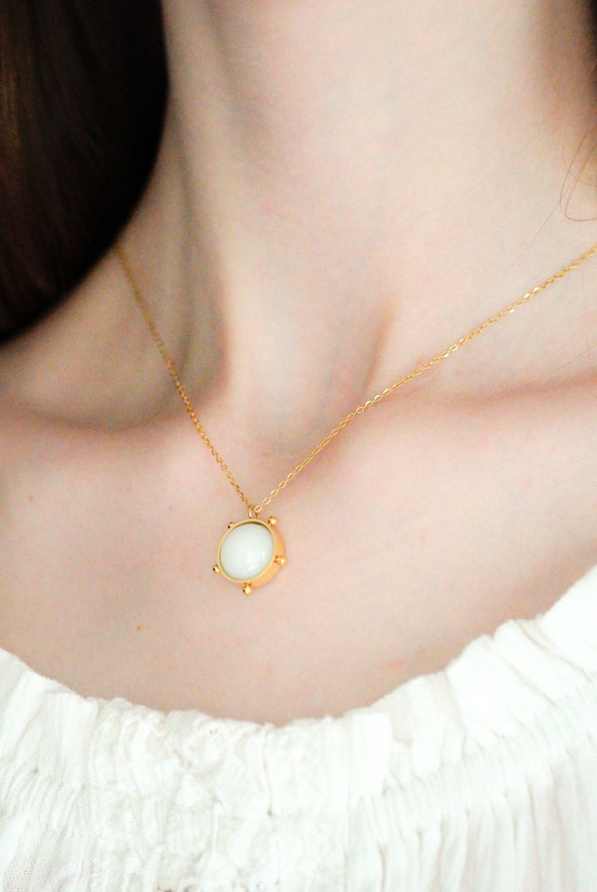 Tuvplāns sievietei, kurai mugurā ir Como Jewelry rotaslietas "Kaklarota - Mēness gaisma" ar apaļu baltu piekariņu, kas kombinēts ar baltu topu bez plecu apakšas. Viņas seja un fons nav redzami.