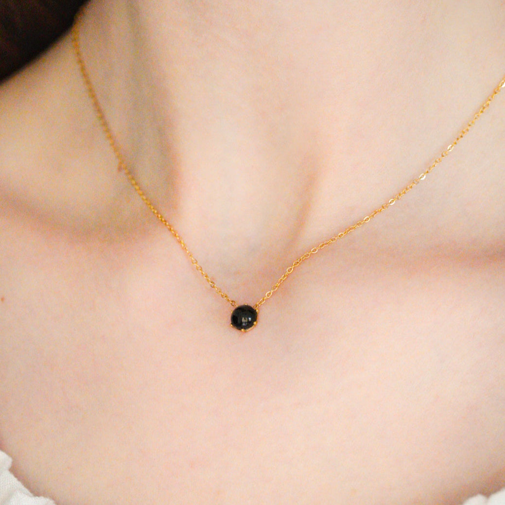 Tuvplāns uz personas kakla, uz kuras kakla valkā Como Jewelry Kaklarota - Pusnakts, ar nelielu melnu akmens piekariņu, kas kombinēts ar baltu rūtainu kreklu.