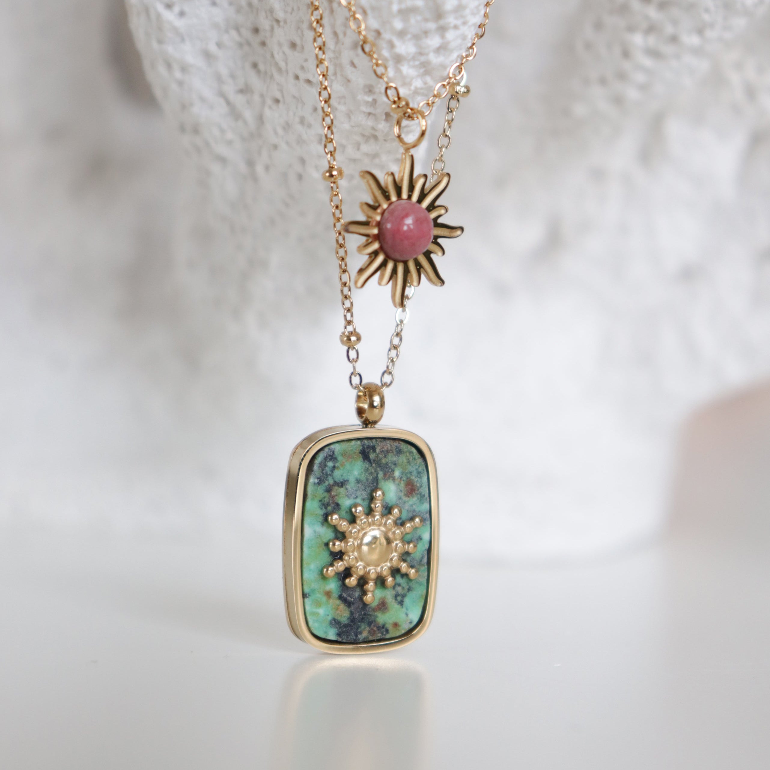 Kaklarota - Saule no Como Jewelry: viena ar taisnstūrveida tirkīza kulonu un zelta saules staru motīvu, otra ar mazu rozā akmeni zelta staros-abas elegantas rotas uz teksturēta balta fona.