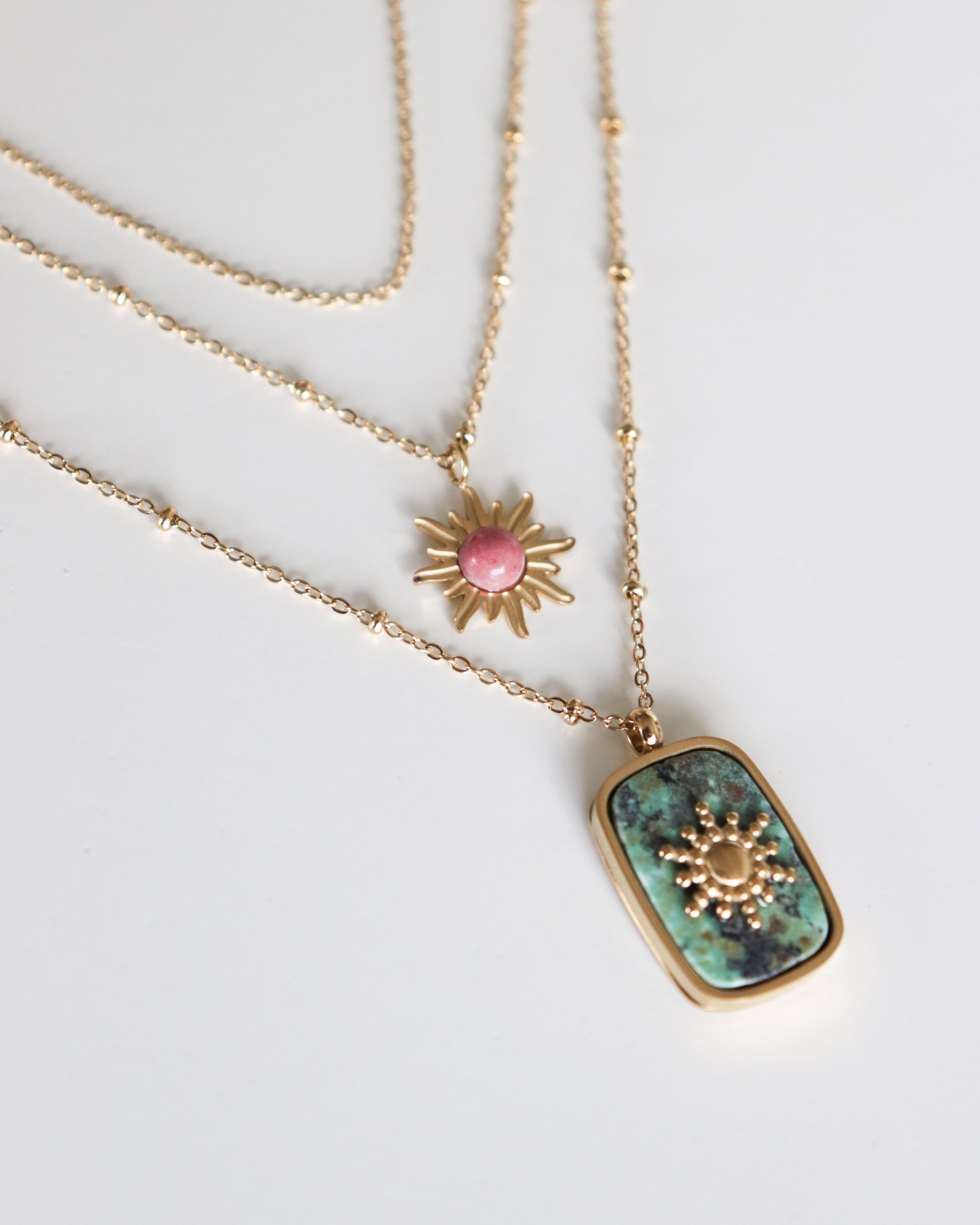 Kaklarota - Saule by Como Jewelry piedāvā divas zeltītas kaklarotas - rozā saules staru piekariņš un taisnstūra formas zaļš akmens ar zelta saules staru. Šī antialerģiska rotaslieta ir mūsdienīga, stilīga un attēlota baltā krāsā.