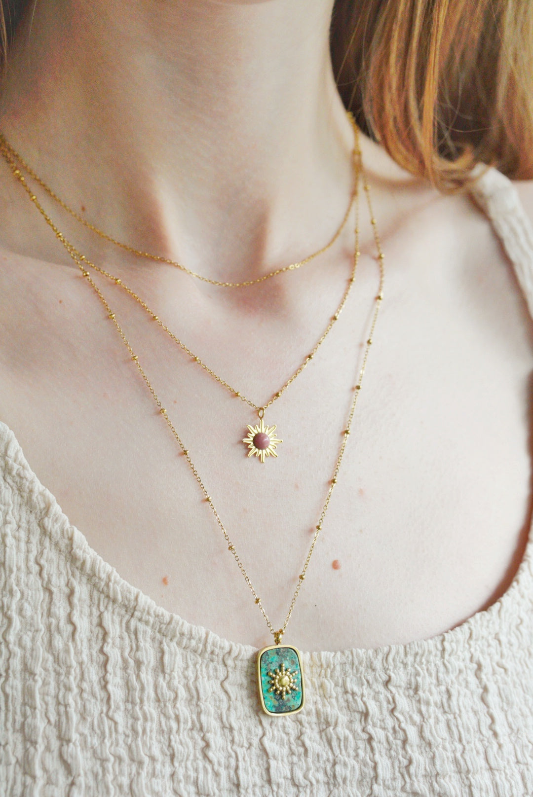 Sieviete valkā krēmkrāsas faktūras topu un Como Jewelry Kaklarota - Saule - modernu zelta kaklarotu ar diviem slāņiem: vienā ir neliels saules kulons, bet otrā - taisnstūra formas tirkīza kulons ar zelta zvaigznīti.