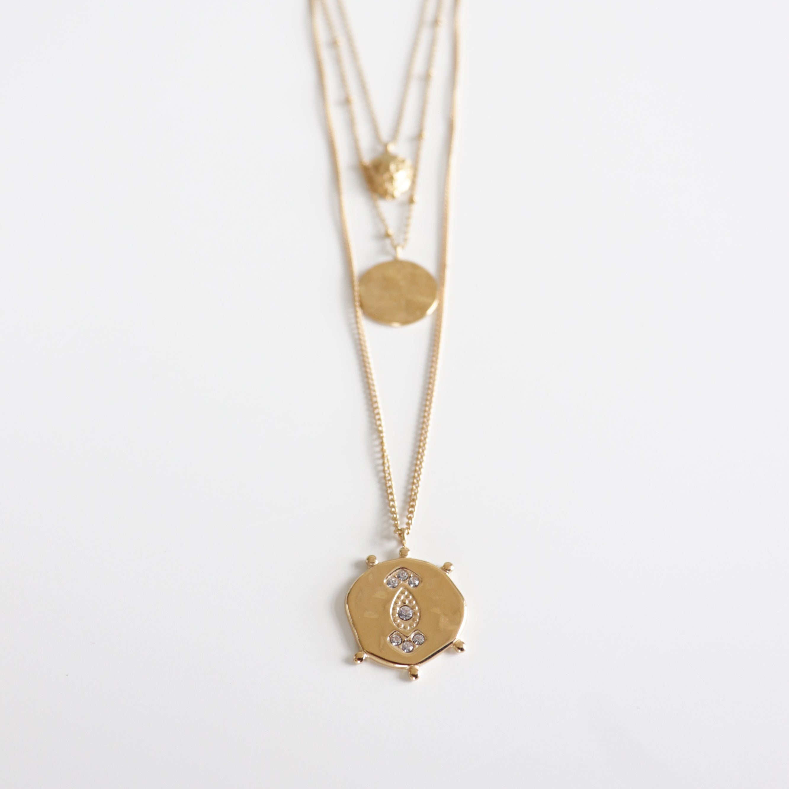 Kaklarotu komplekts Gaismas kārtas no Como Jewelry - trīs zelta kaklarota ar sešstūra kulonu ar akmentiņiem un diviem apaļiem, teksturētiem piekariņiem, modernā stilā uz balta fona.