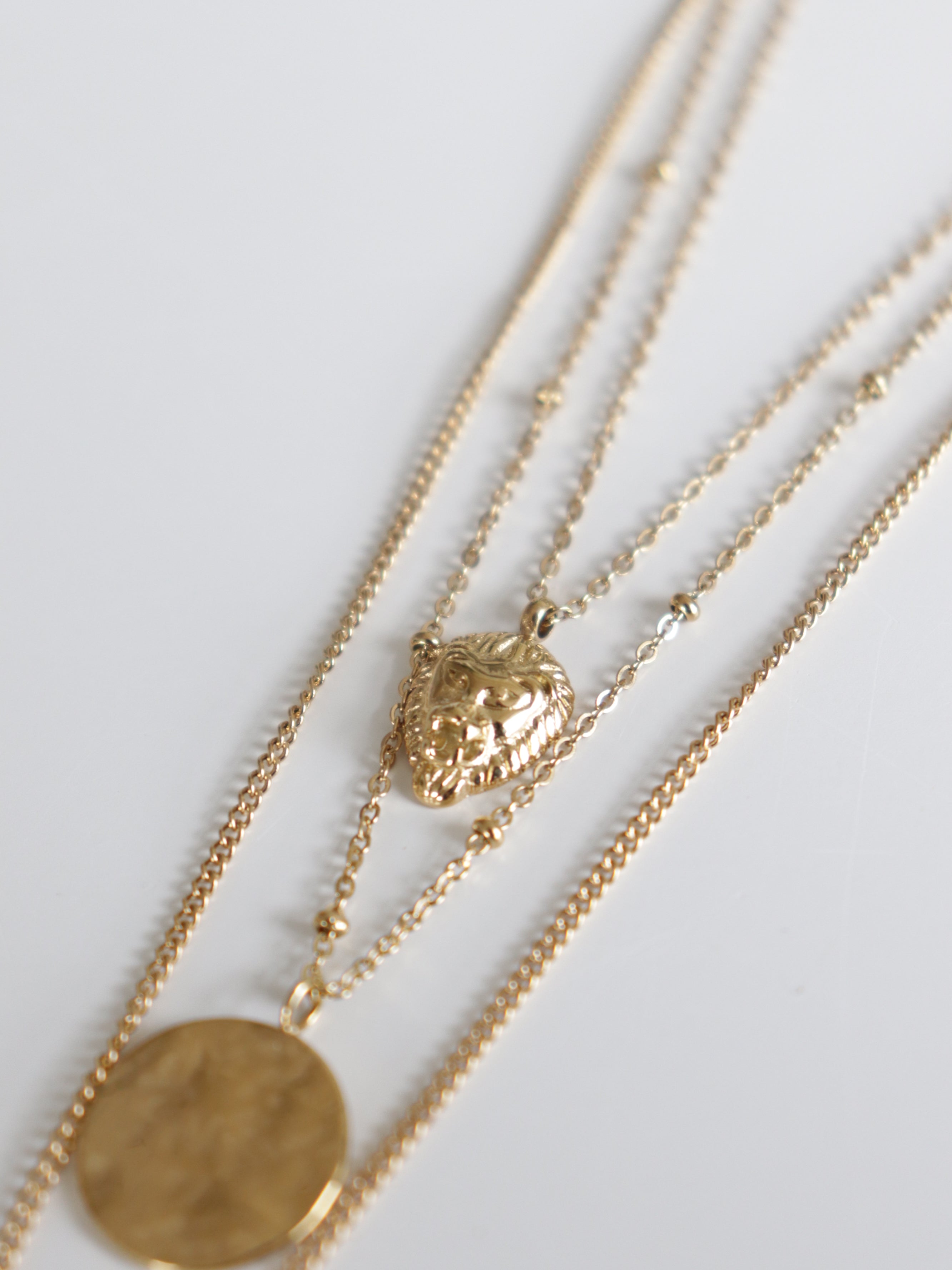 Como Jewelry Kaklarotu komplekts Gaismas kārtas tuvplāns uz baltas virsmas, kurā redzamas daudzslāņainas kaklarotas ar lauvas galvas piekariņu un apaļu monētas formas piekariņu.