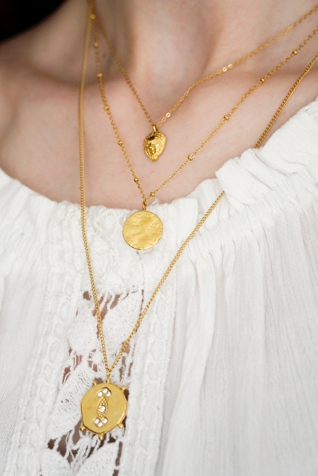 Tuvplānā redzama persona ar baltu faktūru blūzi un Como Jewelry Kaklarotu komplekts Gaismas kārtas: trīs kārtu kaklarotas, katra ar unikālu piekariņu - sirds formas, apaļu un sešstūrainu ar maziem dārgakmeņiem.