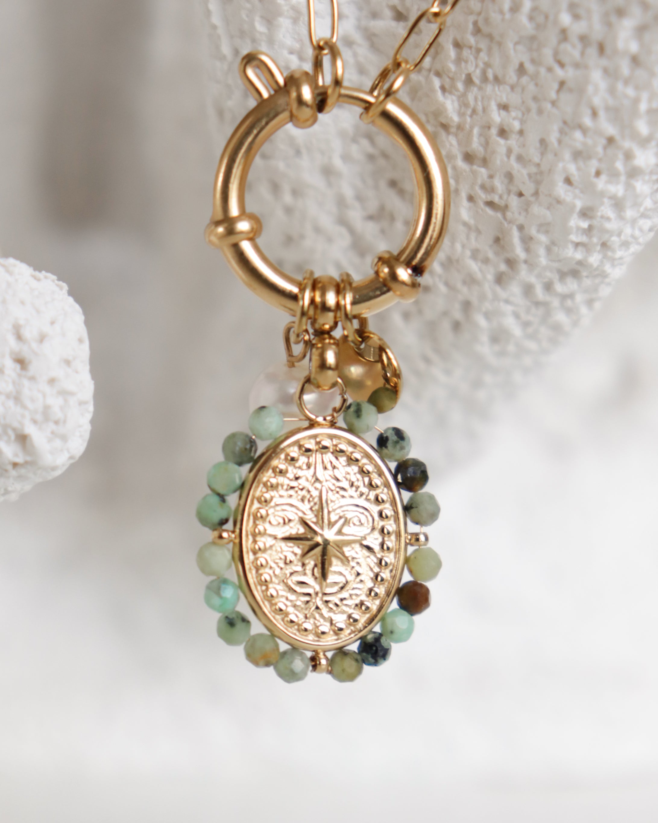 Kaklarota "Vēja roze" no Como Jewelry ar zelta kulonu, reljefu zvaigznes motīvu un zaļām un brūnām pērlītēm, eleganti karājas uz teksturēta gredzena; antialerģiska rotaslieta sievietēm.