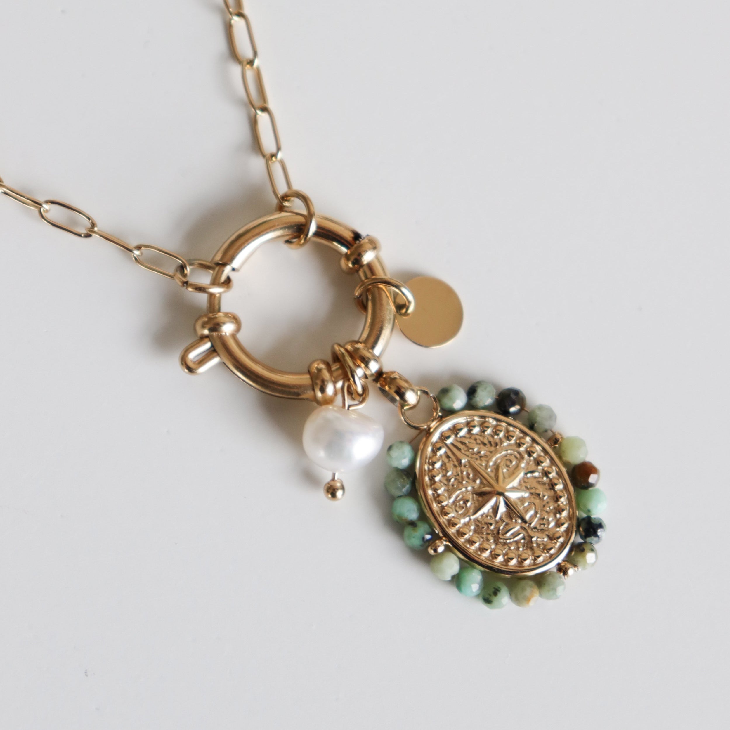 Kaklarota - Vēja roze no Como Jewelry: moderna zelta ķēde ar apaļu aizdari, teksturētu medaļas piekariņu, zaļām pērlītēm, baltu pērli un nelielu zelta disku uz balta fona.