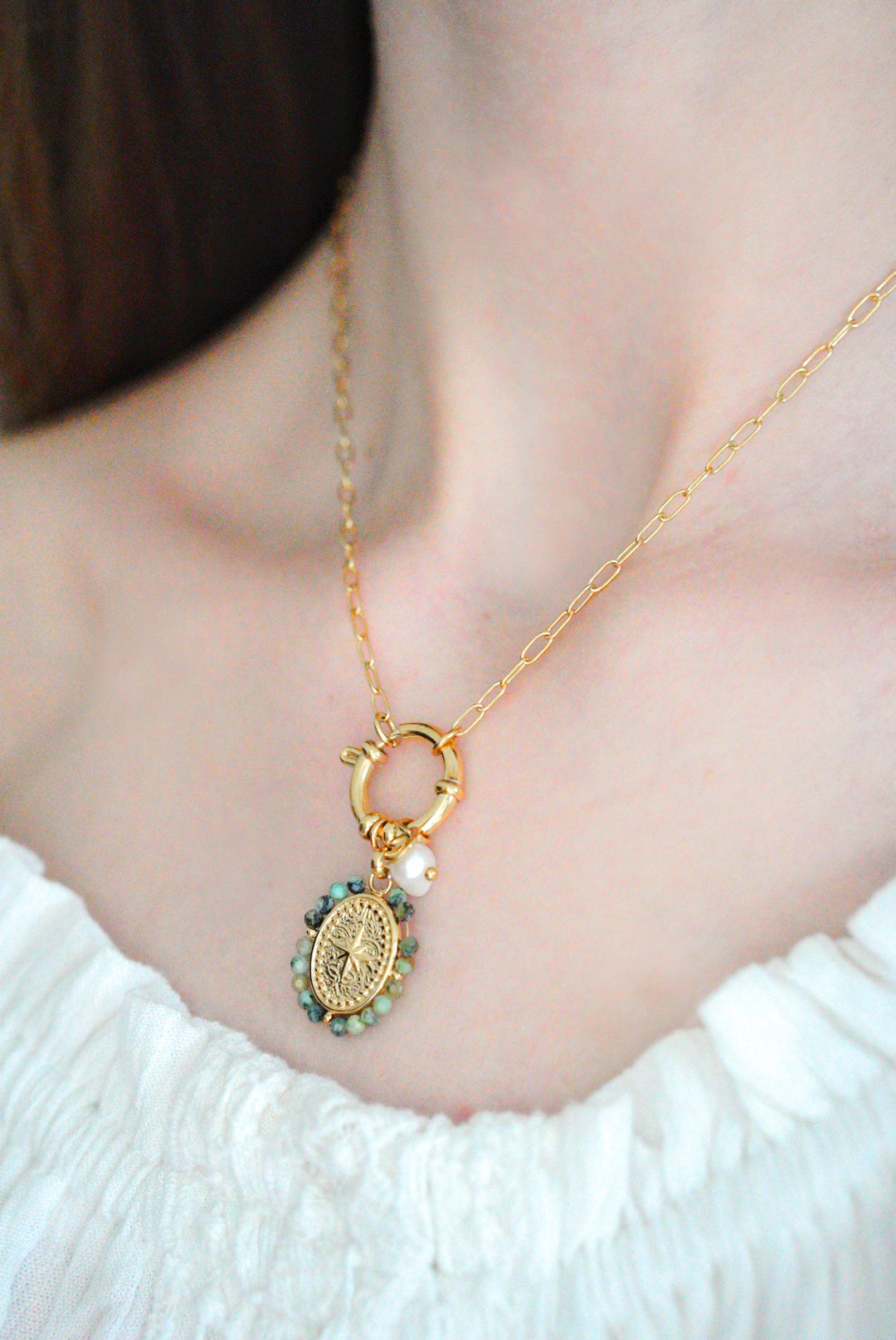 Kaklarota - Vēja roze" by Como Jewelry-zelta kaklarota ar apaļu kulonu, zaļām pērlītēm un pērļu akcentu, valkāta sievietes ar baltiem, plecus atsedzošiem topu.