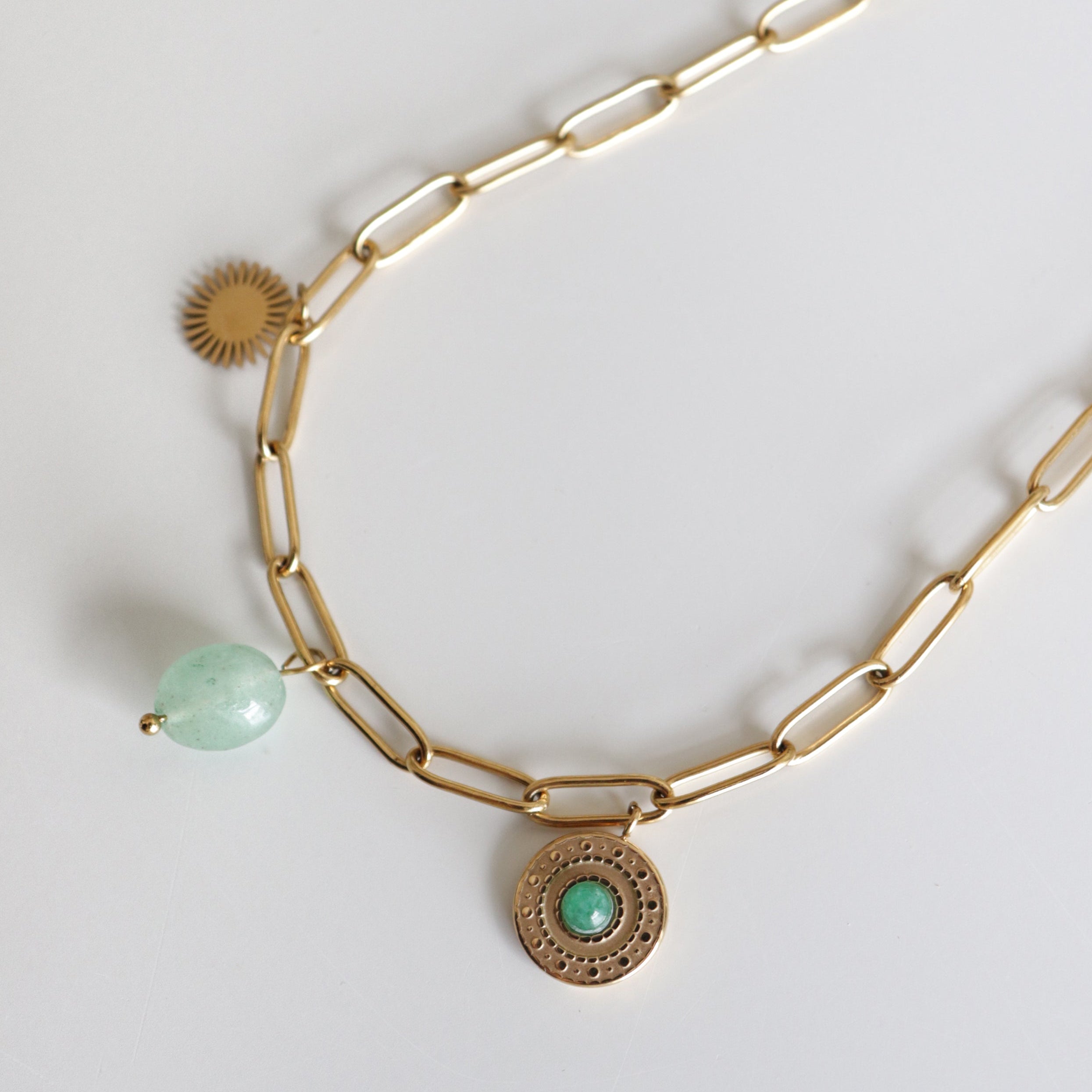 Kaklarota "Zaļās saules ceļš" no Como Jewelry - zelta kaklarota ar trīs piekariņiem: saules staru, zaļu akmens bumbiņu un medaljonu ar zaļu centru, moderna dizaina rotājums uz balta fona.
