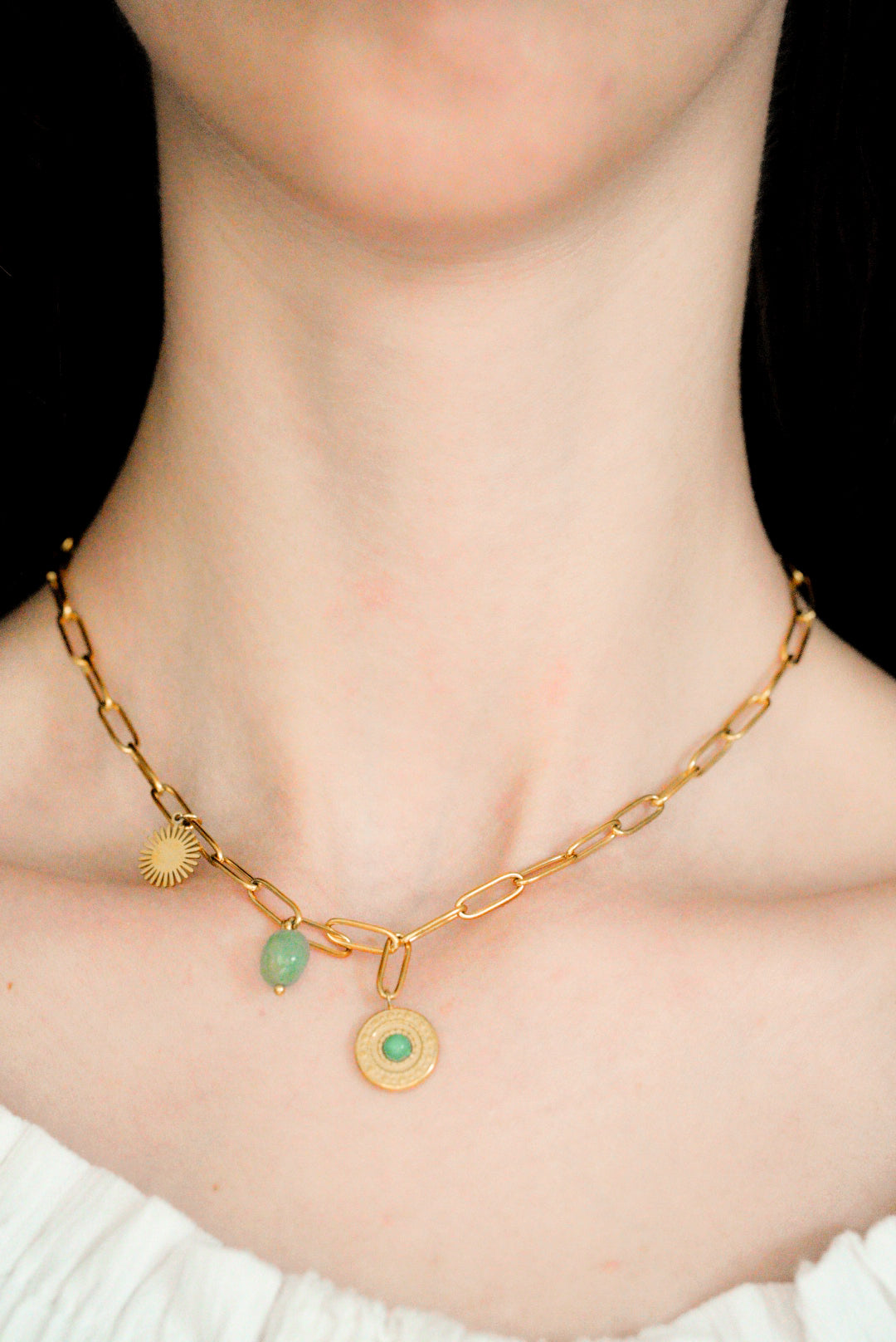 Tuvplāns uz personas kakla, kurai ir Como Jewelry "Kaklarota - Zaļās saules ceļš", mūsdienīga dizaina, antialerģiska kaklarota ar trim zelta piekariņiem - sauli, zaļu akmeni un apaļu medaljonu -, kas ir savienota ar baltu atkailinātu kreklu.