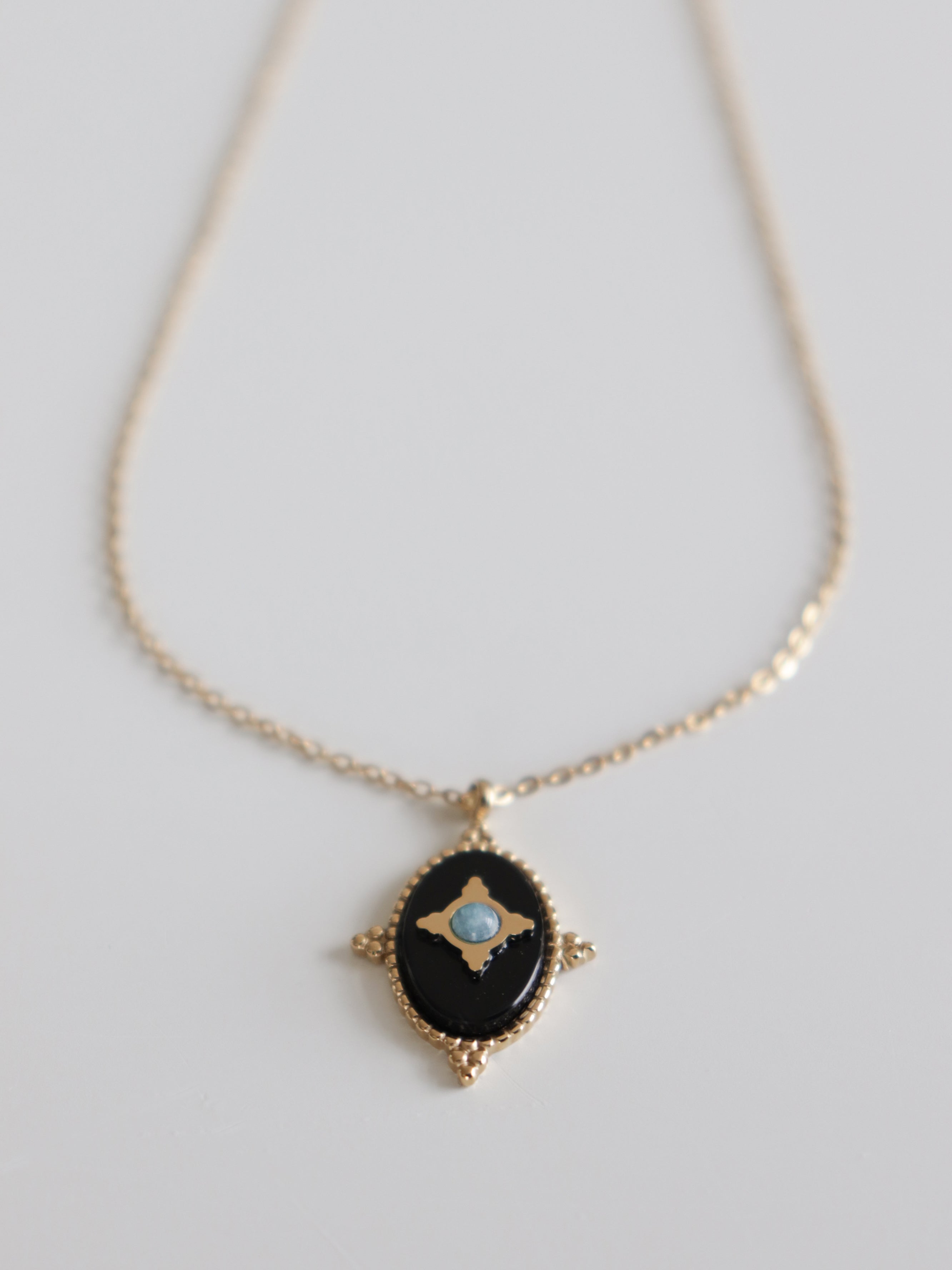 Mūsdienīga dizaina zelta kaklarota sievietēm - Como Jewelry "Kaklarota - Pusnakts talismans" ar ovālu melnu kulonu un mazu zilakmeņu centru, izstādīta uz balta fona.