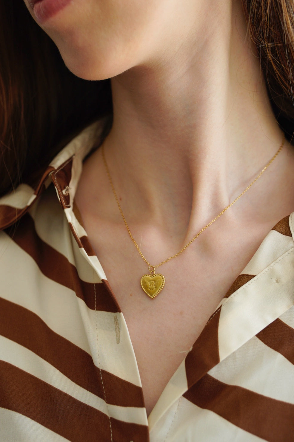 Sieviete modelē Como Jewelry "Kaklarota - Sirds" - zelta piekariņu sirds formā. Uzmanības centrā ir viņas kakls un ūdensizturīgā kaklarota, un viņas seja ir daļēji ārpus kadra.