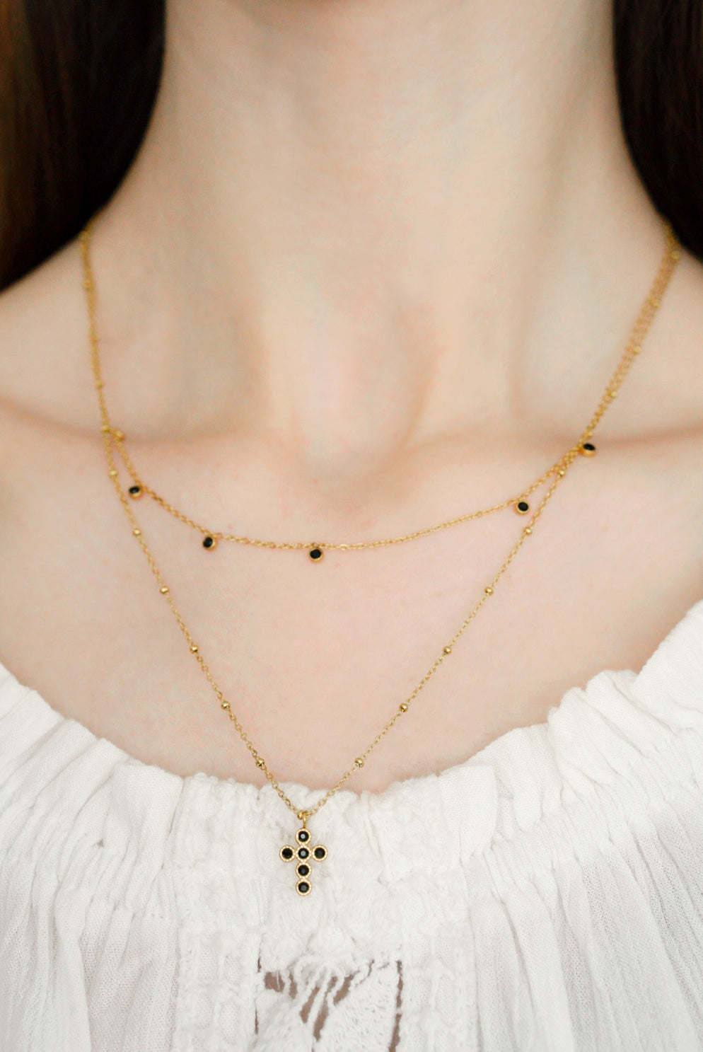 Sieviete valkā Como Jewelry "Kaklarota - Mēness ēna" - daudzslāņainu kaklarotu ar maziem melnu akmentiņu akcentiem. Garākajai ķēdītei ir krusta formas melna akmens piekariņš, kas apvienots ar baltu fakturētu blūzi elegantam, pārlaicīgam izskatam.